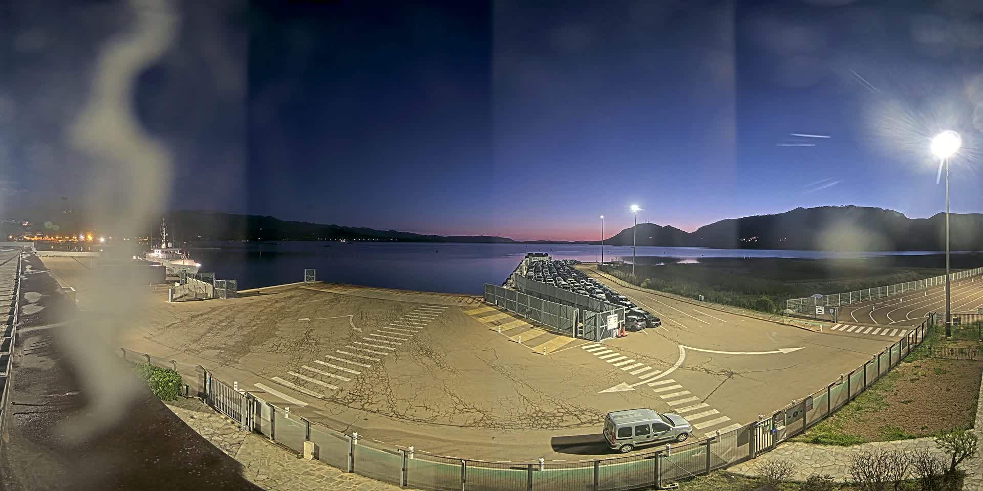An empty port at dusk, with a row of cars parked near the water, under a clear, twilight sky.