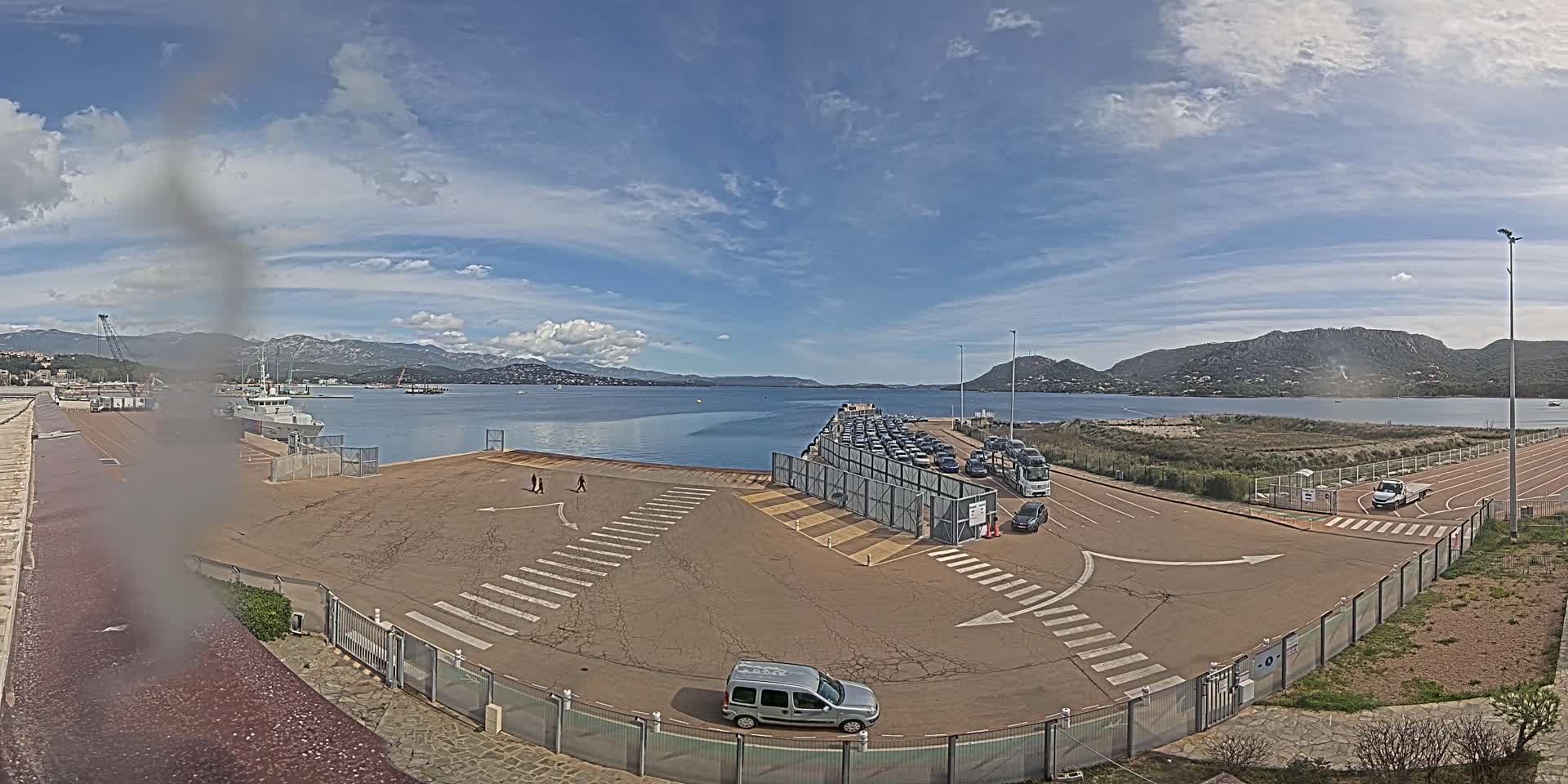 A ferry terminal with numerous cars waiting to board, under a partly cloudy sky, overlooks a calm body of water and mountains in the distance.