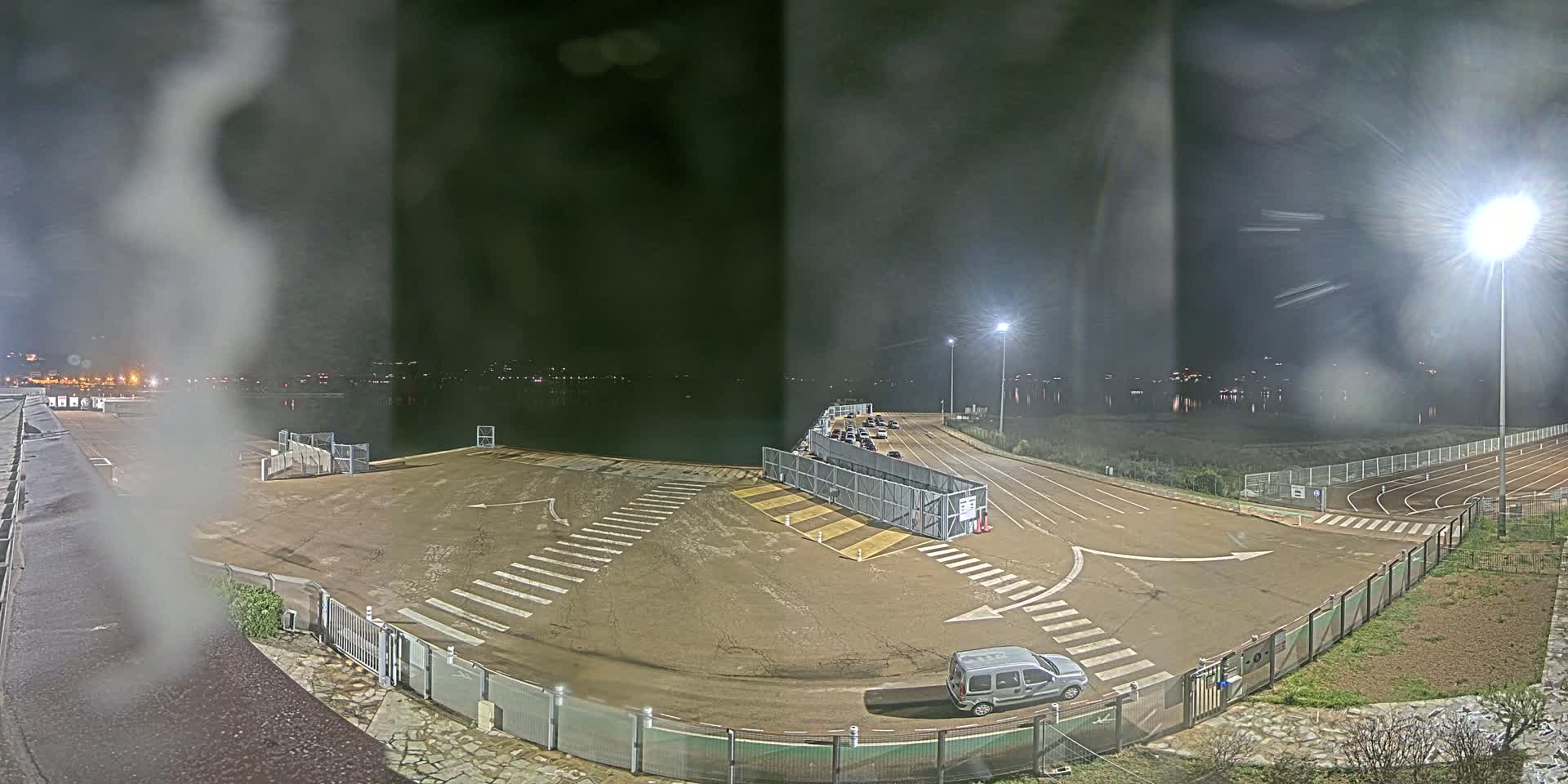 A nighttime view of an empty parking lot near a body of water, with a single vehicle parked and some faint lights in the distance, under slightly hazy conditions.