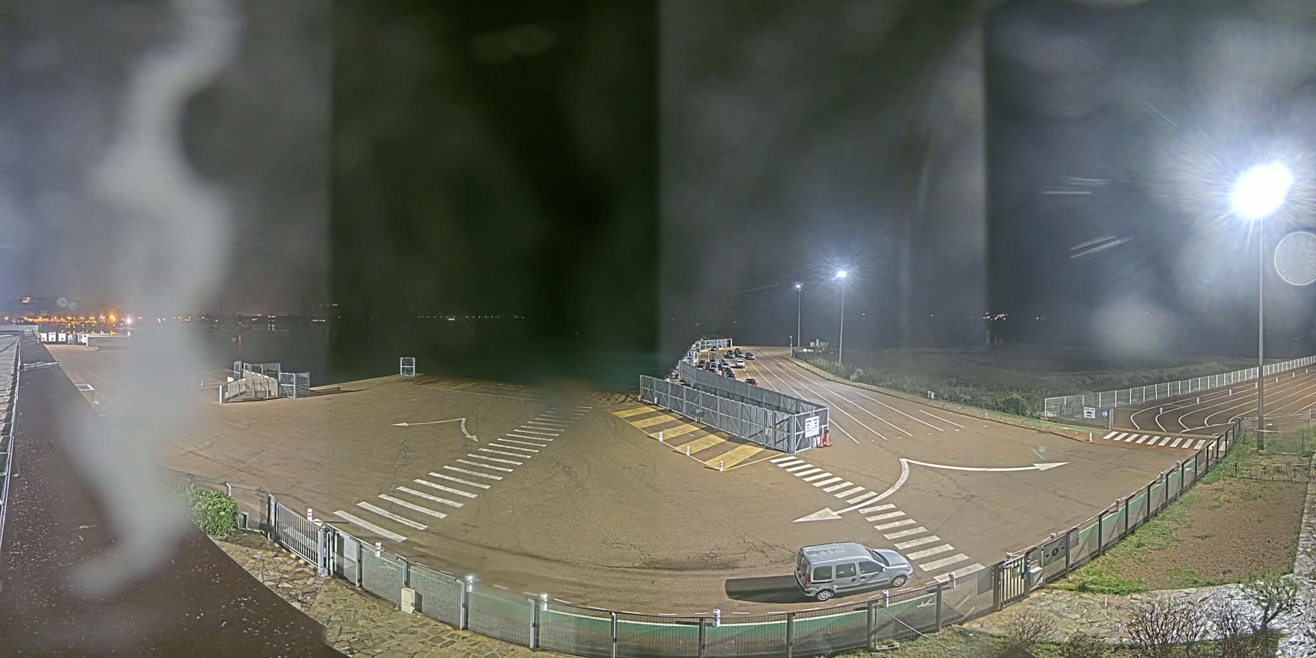 A nighttime, rain-streaked view of an empty parking lot and roadway, with a few vehicles and a gated area, near a body of water under bright lights.