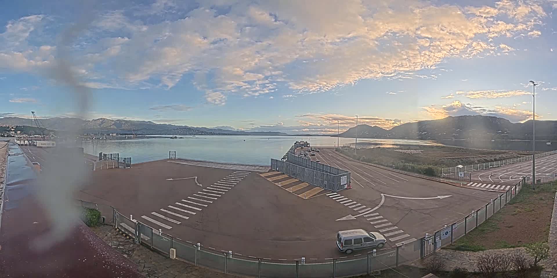 A ferry terminal with a few cars, under a partly cloudy sky, overlooks calm water and distant mountains.