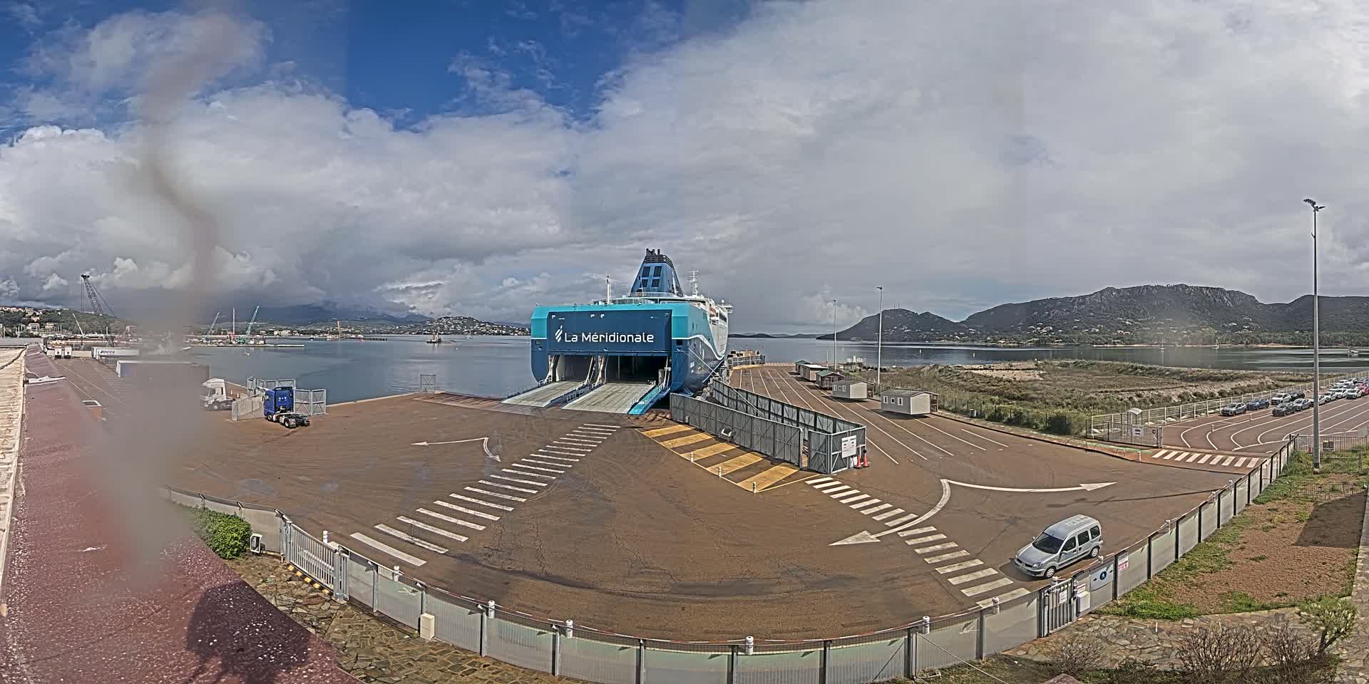 A large ferry is docked at a port under a partly cloudy sky, with several vehicles parked nearby and mountains visible in the distance.