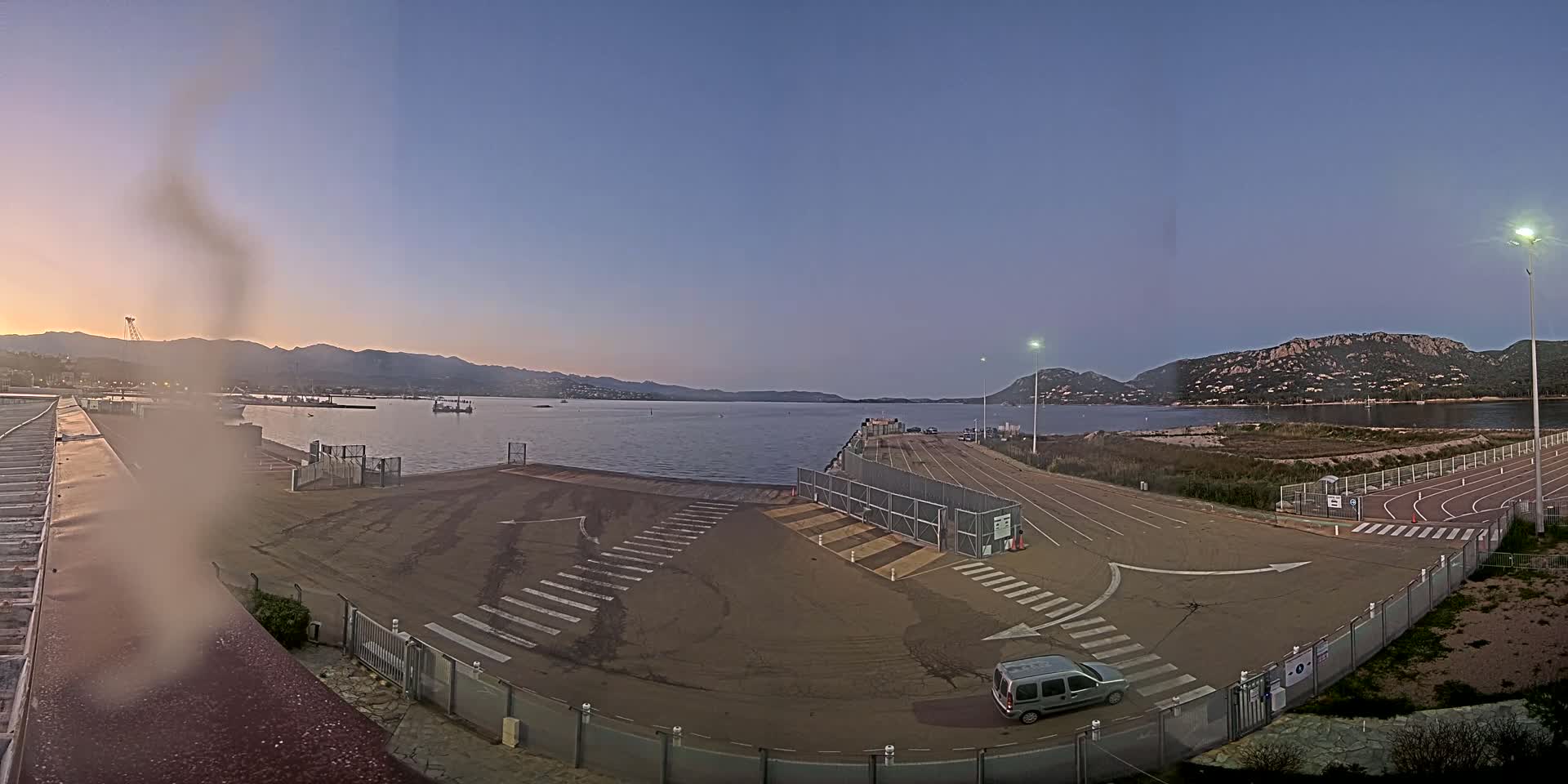 A mostly empty ferry terminal with a calm body of water and mountains in the background under a twilight sky.