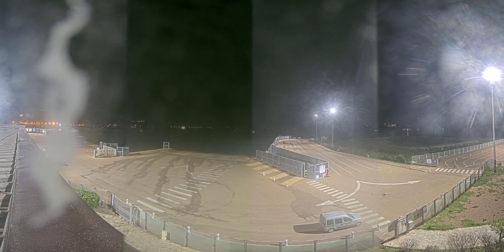 A nighttime view of an empty parking lot and roadway, possibly near a waterfront, with a single vehicle present and some fog or mist in the air.