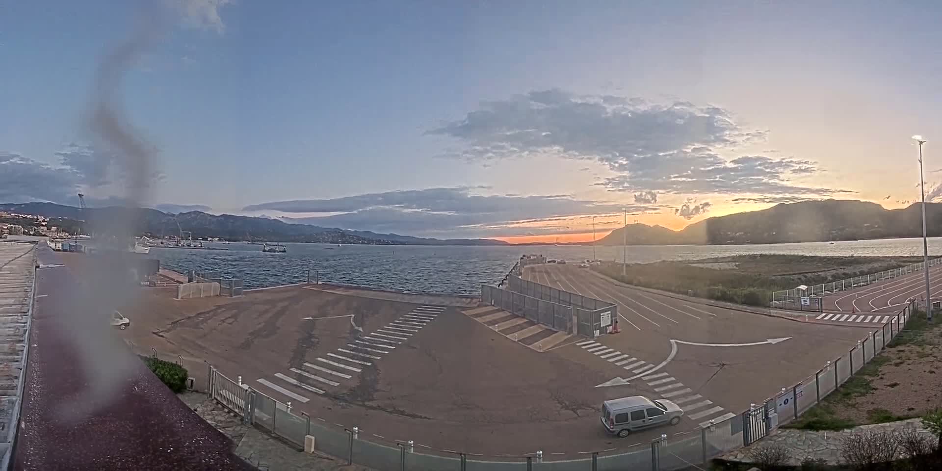 A waterfront area with a paved road, crosswalks, and a few vehicles is seen during sunset under a partly cloudy sky, with calm water and mountains in the background.