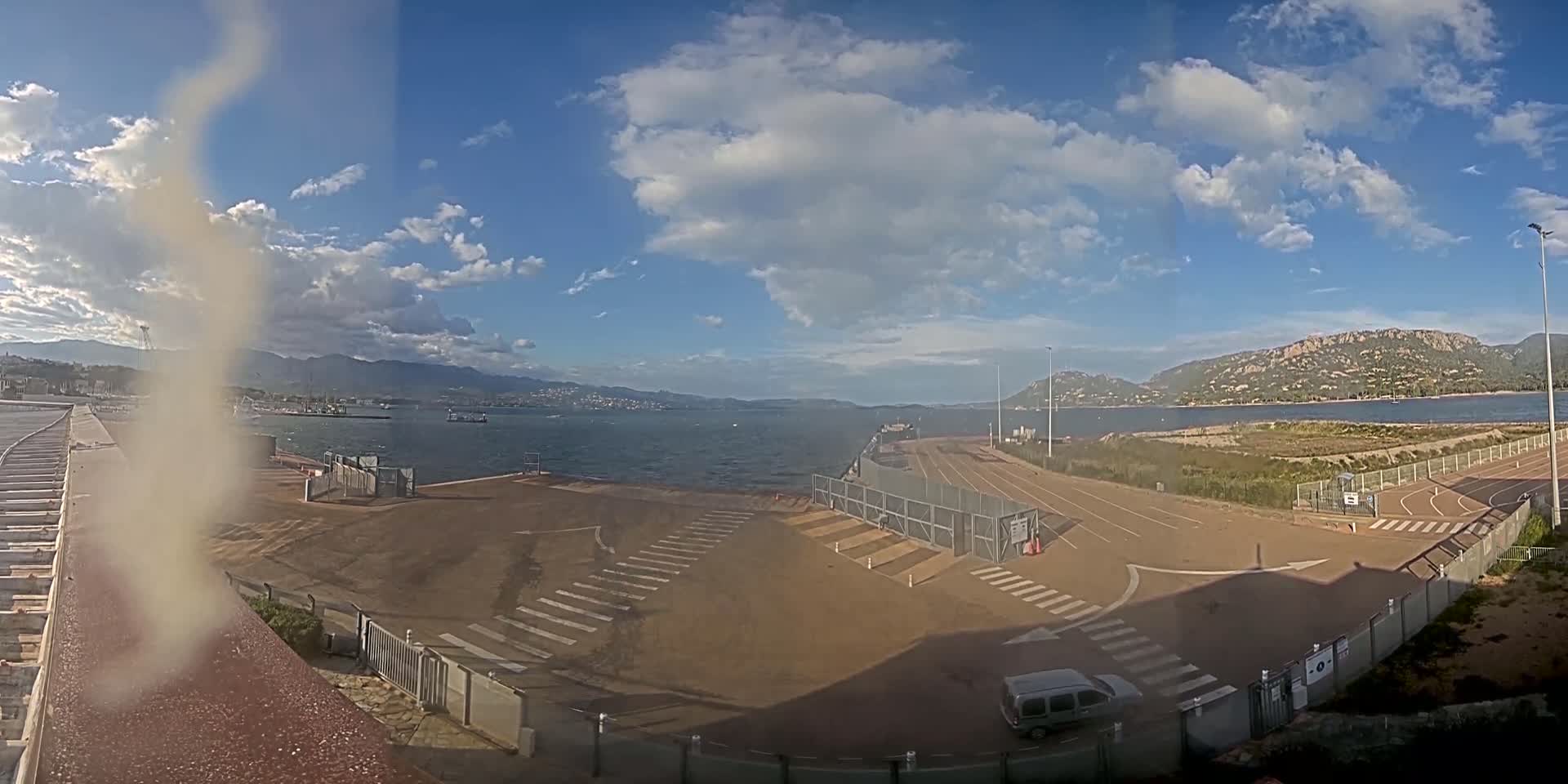 A mostly sunny day with some clouds overlooks a waterfront area with a paved lot, fences, and a single vehicle parked near the water.