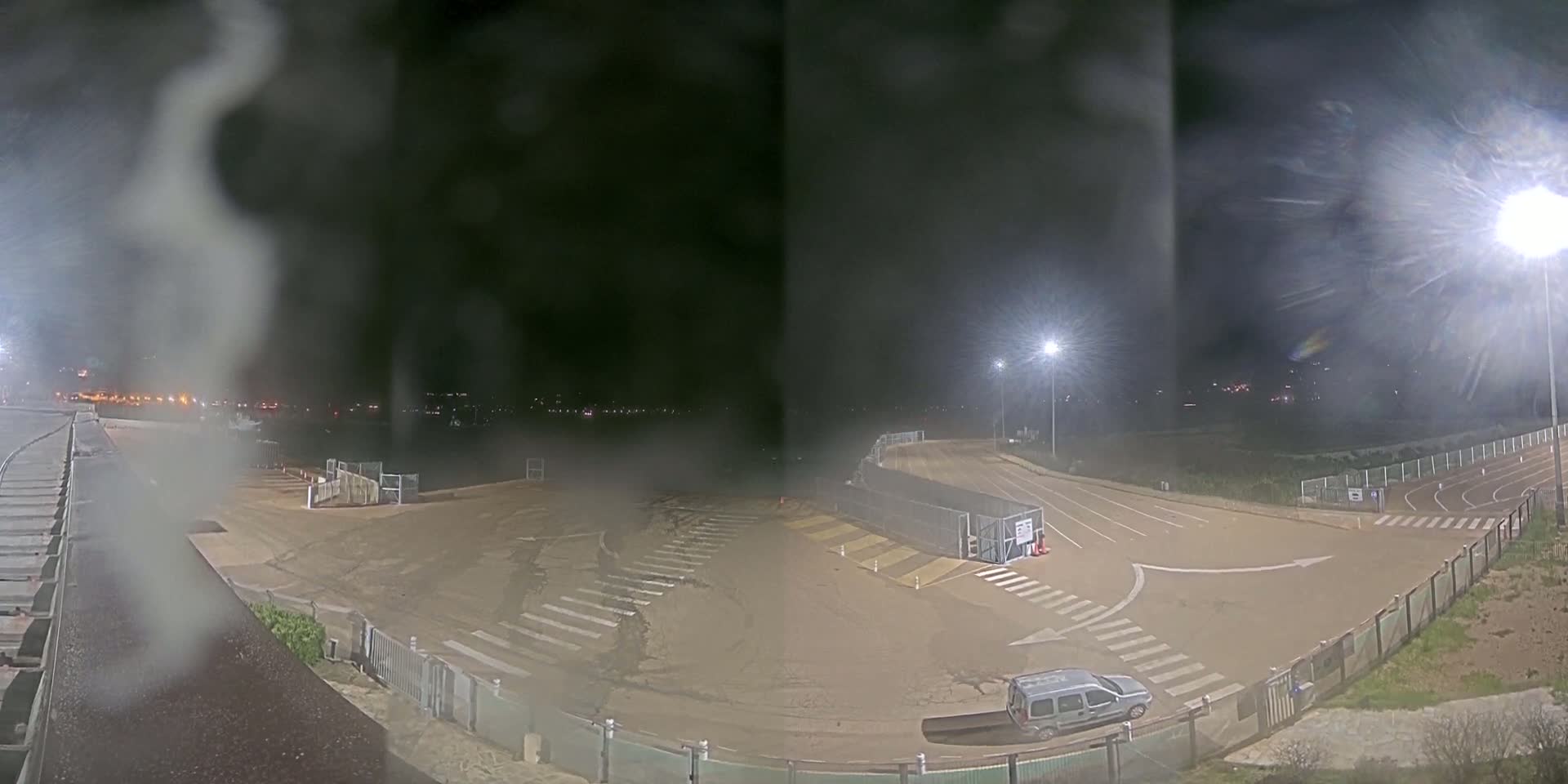 A nighttime, hazy view of an empty paved area with a vehicle, surrounded by fences and lighting.