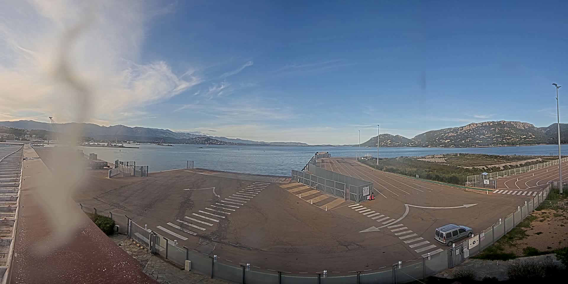 An empty ferry terminal with crosswalks and a single car, overlooks a calm body of water surrounded by mountains under a mostly clear sky.