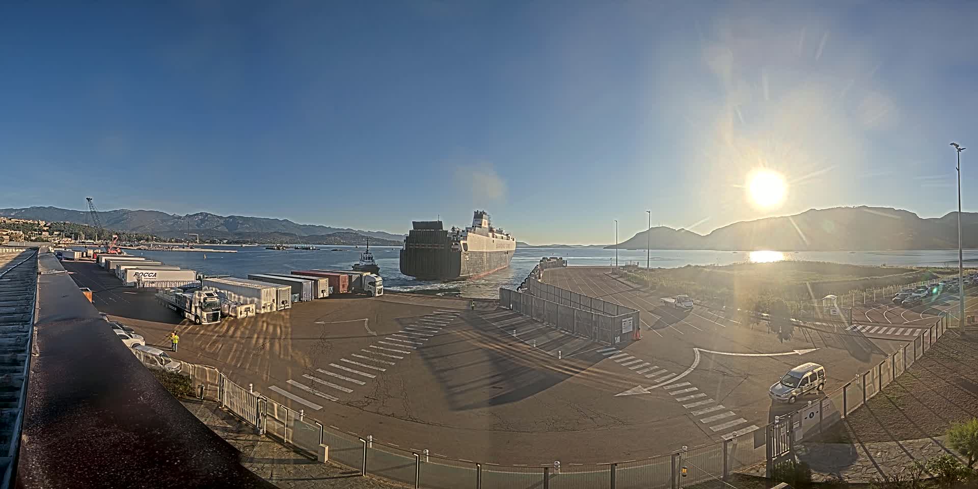 A large cargo ship is docked at a port on a sunny day, with mountains and calm water visible in the background, and several trucks and cars parked nearby.