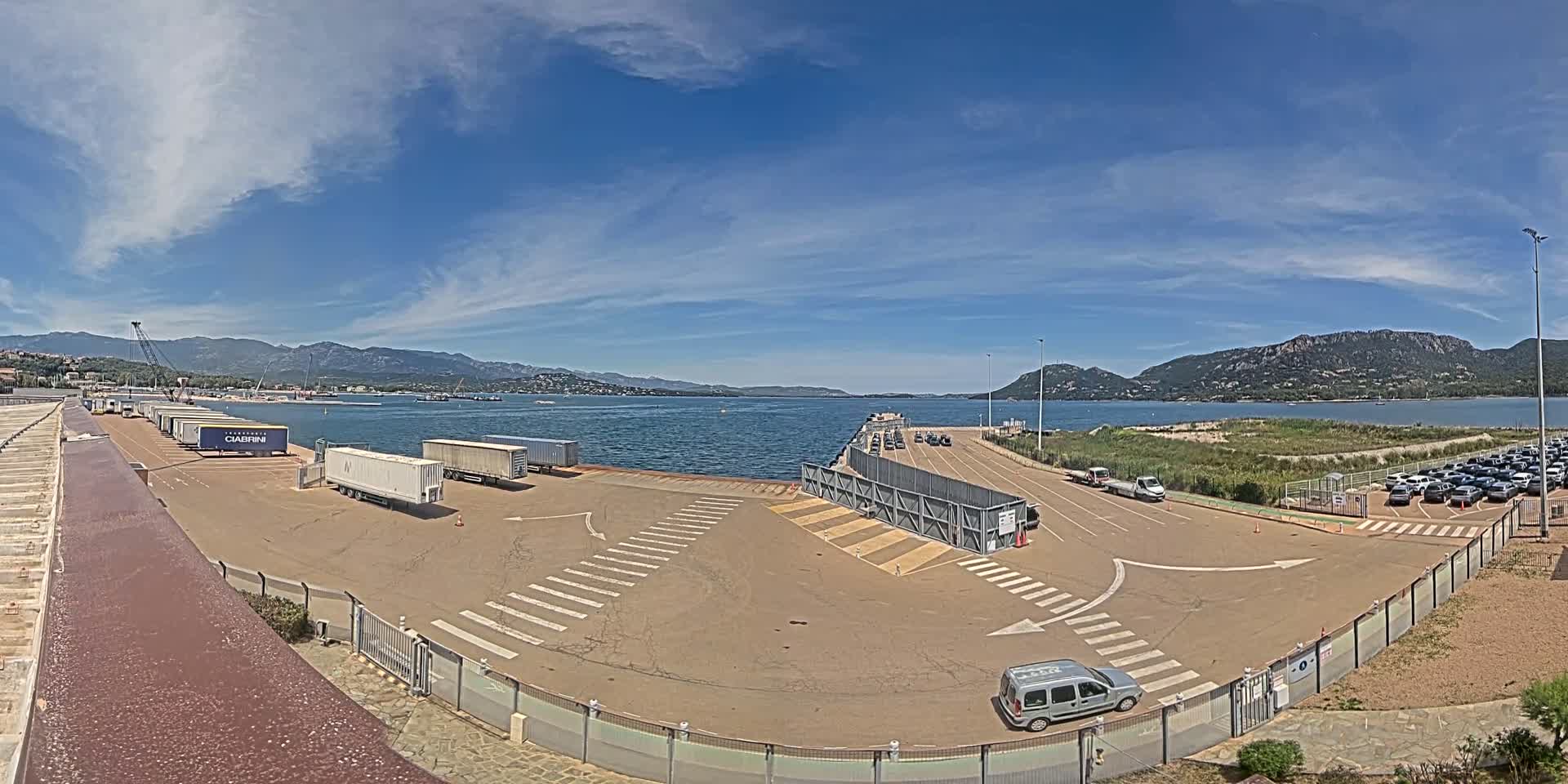 A ferry terminal with several trailers and cars, under a mostly sunny sky with some clouds, overlooks a calm body of water surrounded by mountains.