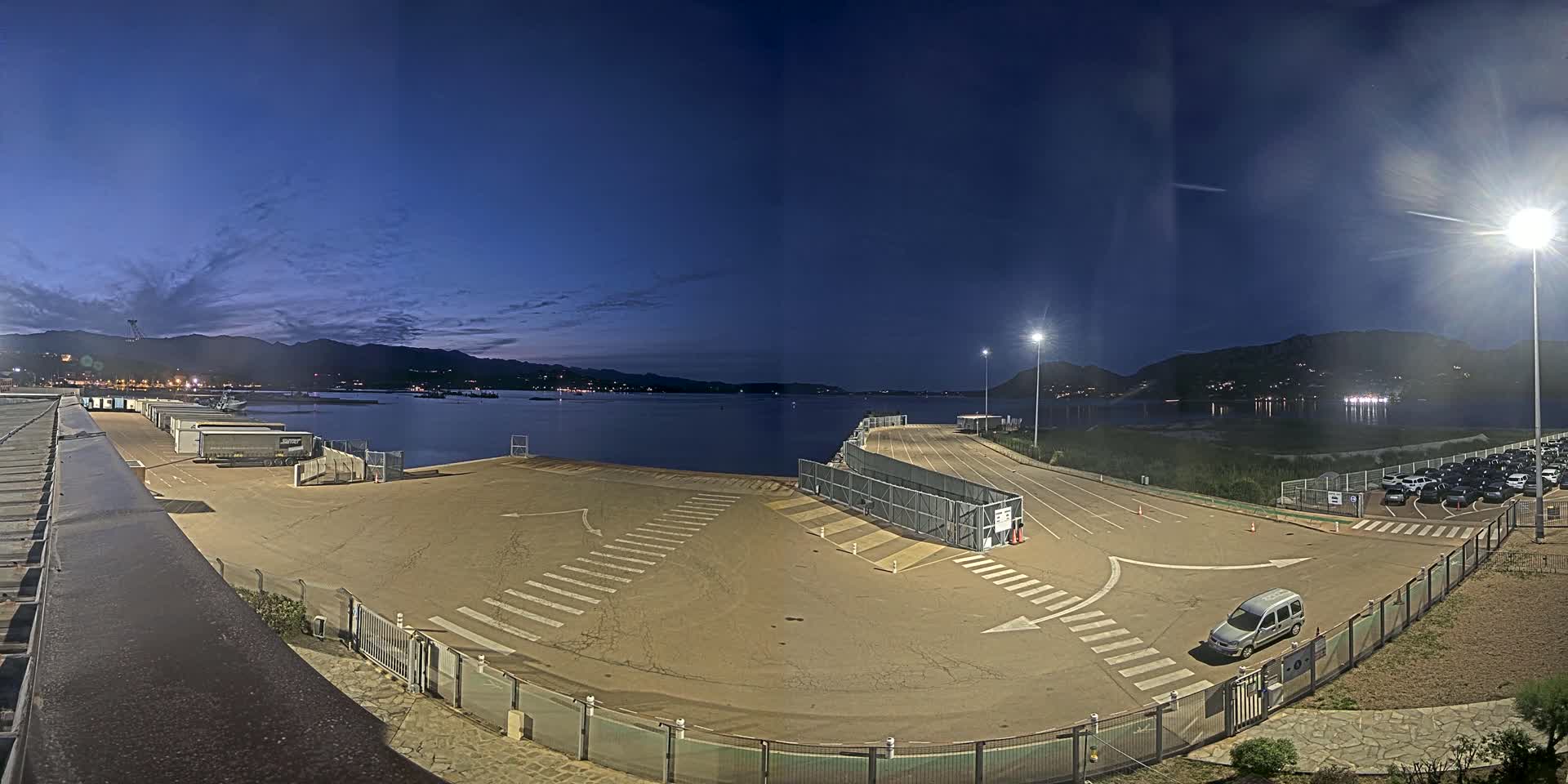 An empty ferry terminal at night, with a calm body of water and mountains in the background under a twilight sky.