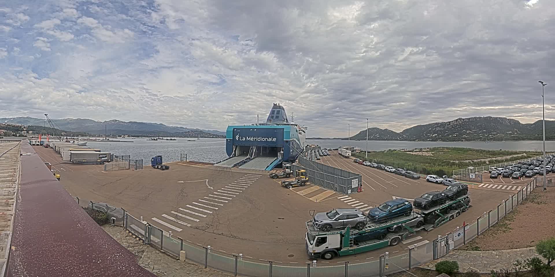 A large ferry is docked at a port under a cloudy sky, with numerous cars parked nearby and being loaded onto a car carrier.