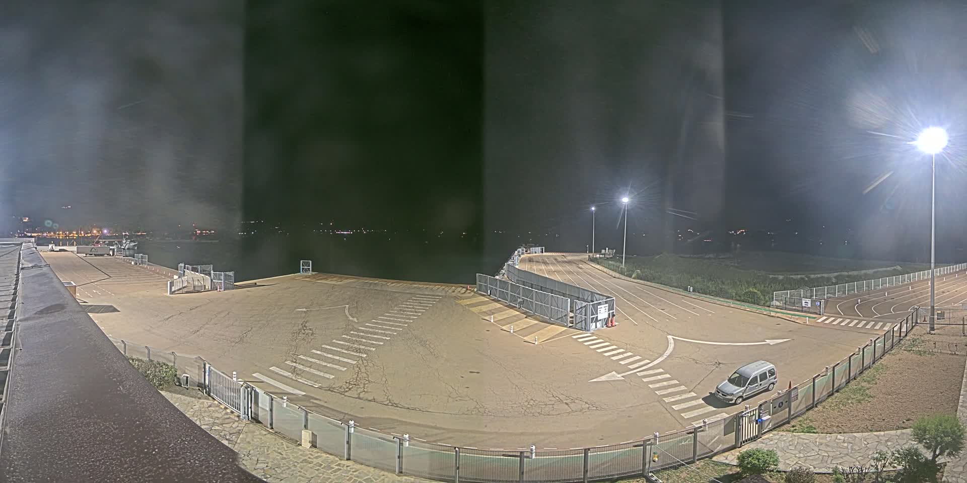 A mostly empty, paved area with crosswalks and fencing at night, illuminated by artificial lights, overlooks a dark body of water.