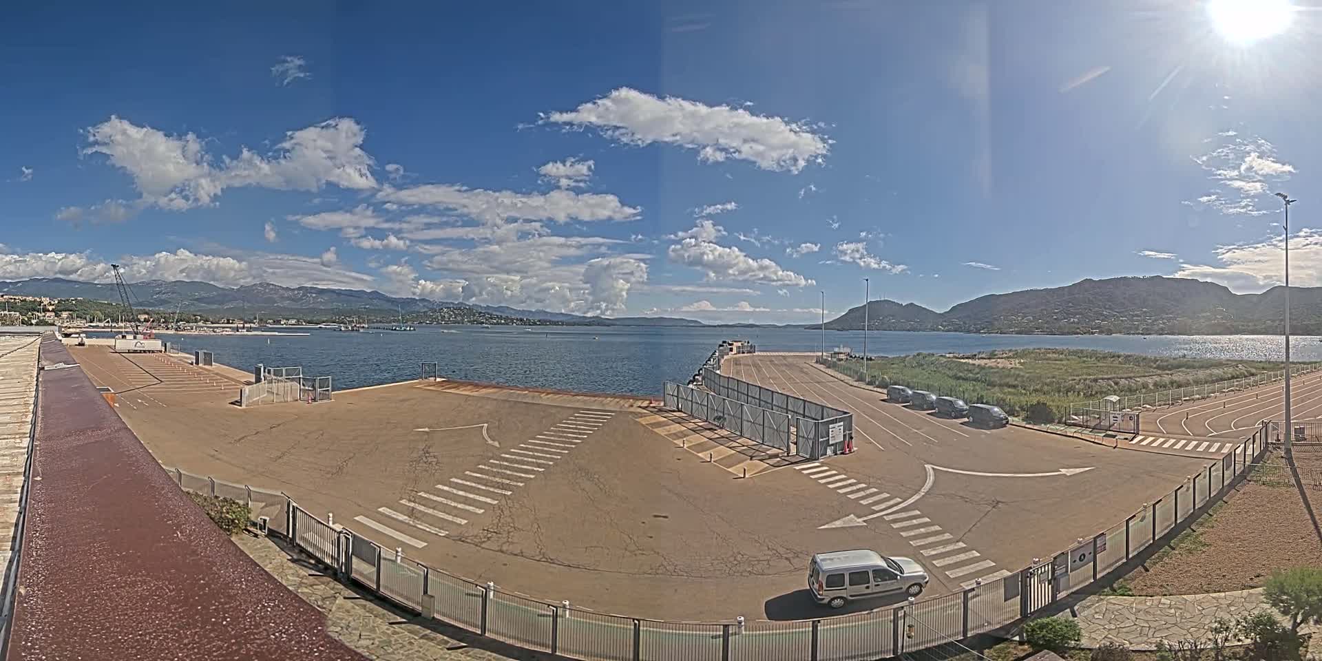 A mostly sunny day at a port with a few cars parked, a mostly empty parking lot, and mountains in the distance.