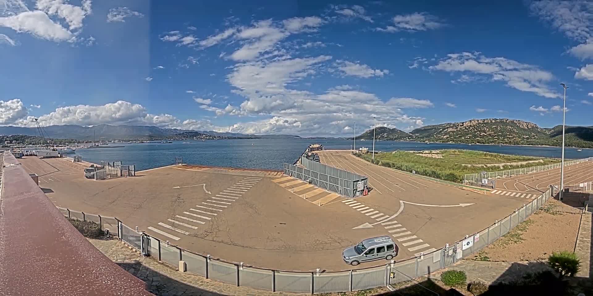 A mostly sunny day overlooks a port area with a single vehicle parked in a paved lot near a body of water, mountains in the background.