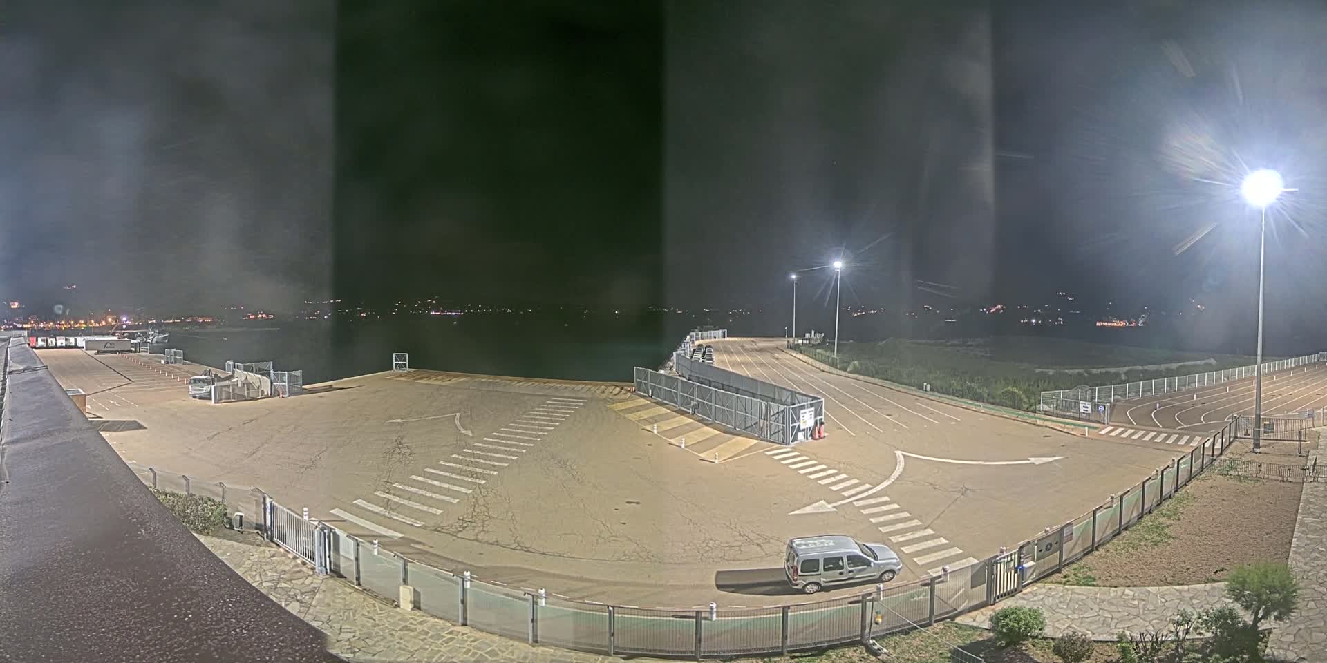 A nighttime view of an empty, paved lot near a body of water, illuminated by several bright lights, with a single vehicle parked.