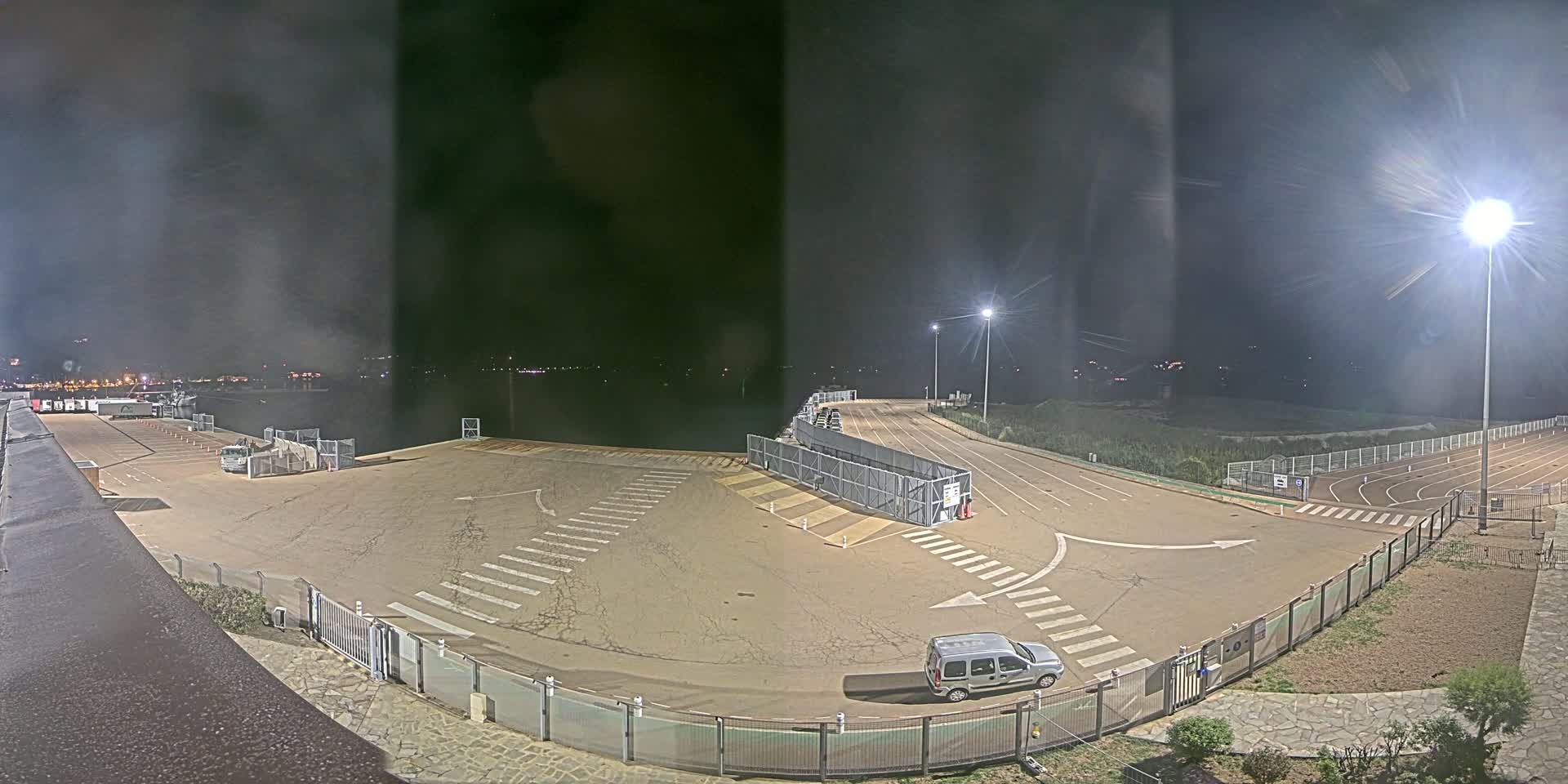 An empty, well-lit nighttime parking lot near a body of water, bordered by fencing and a running track.