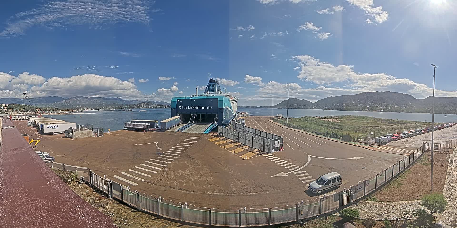 A large ferry is docked at a port on a partly cloudy day, with numerous vehicles parked nearby and mountains visible in the distance.
