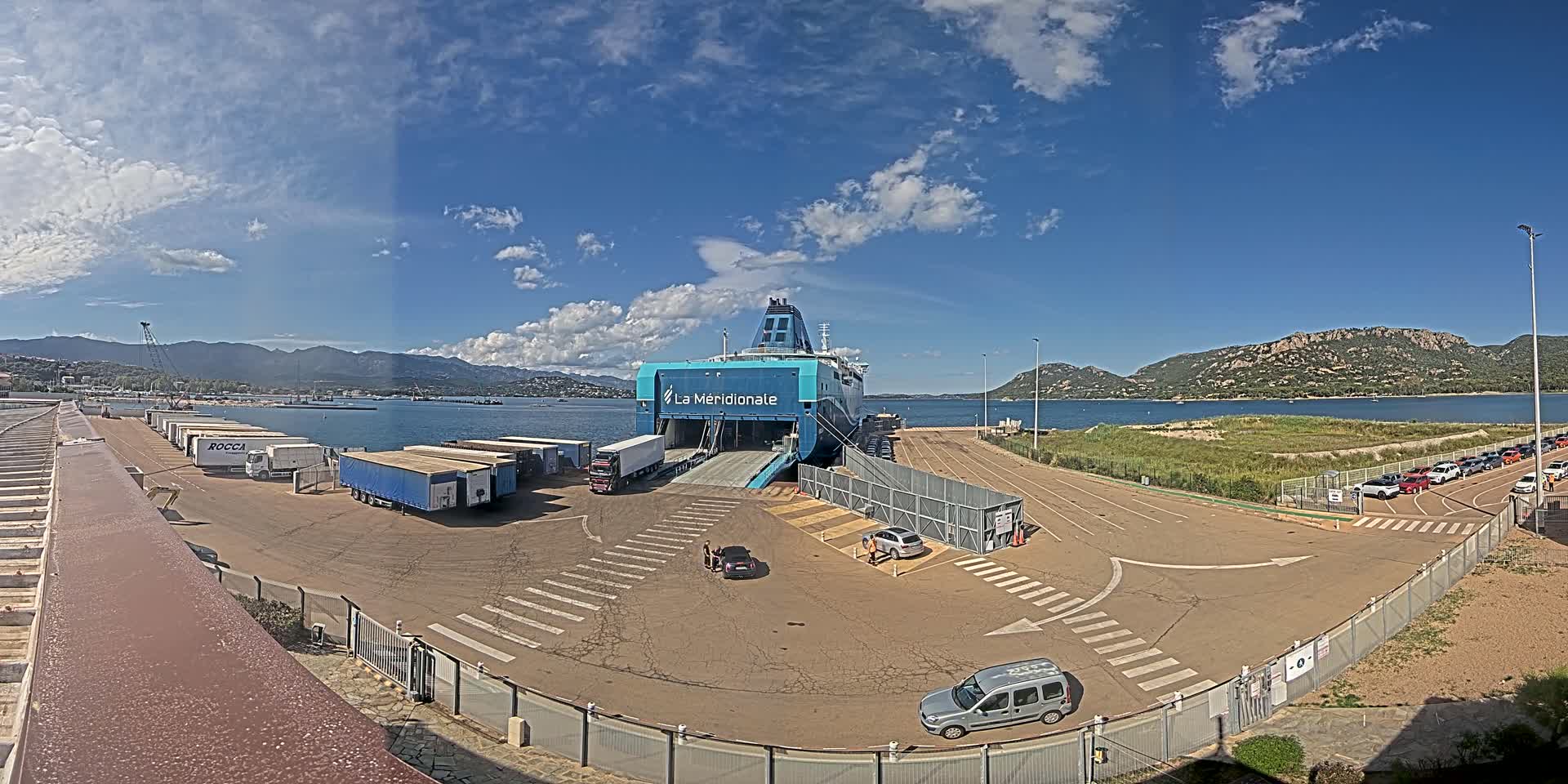 A ferry is docked at a port on a sunny day, with several trucks and cars parked nearby, mountains in the background, and calm waters.