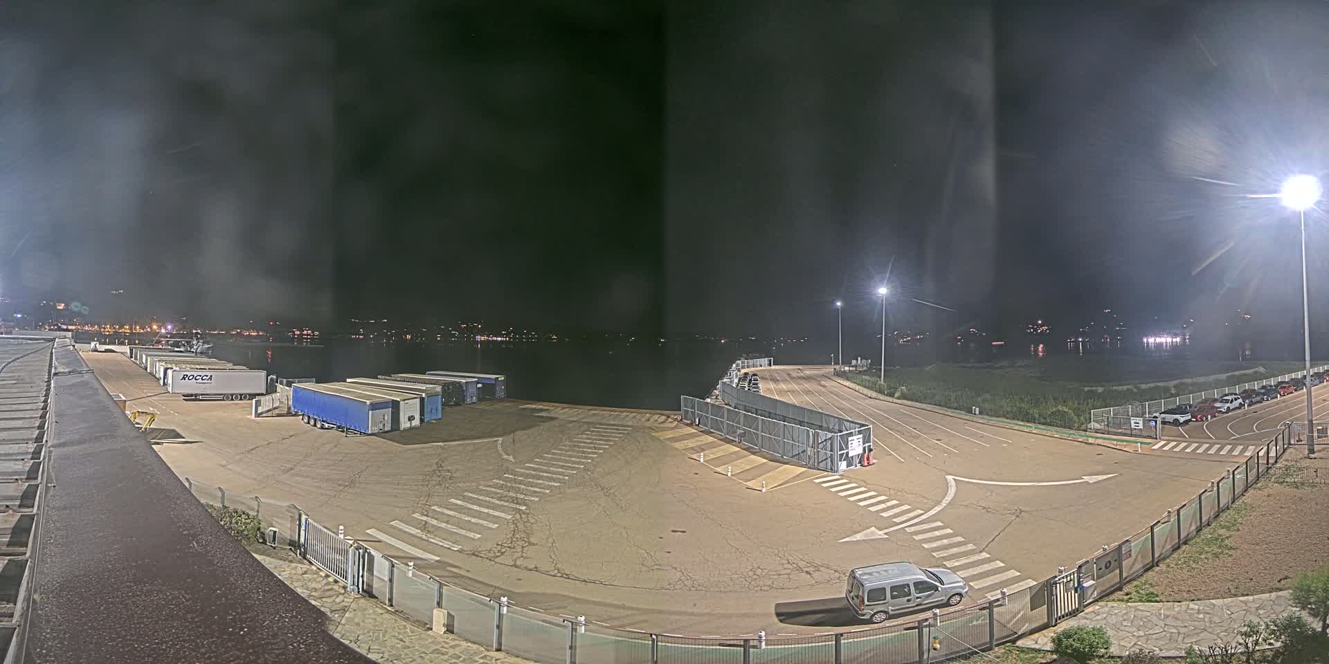 At night, a mostly empty port area with several parked semi-trucks and a few cars is illuminated by bright lights, overlooking calm water with distant lights on the opposite shore.