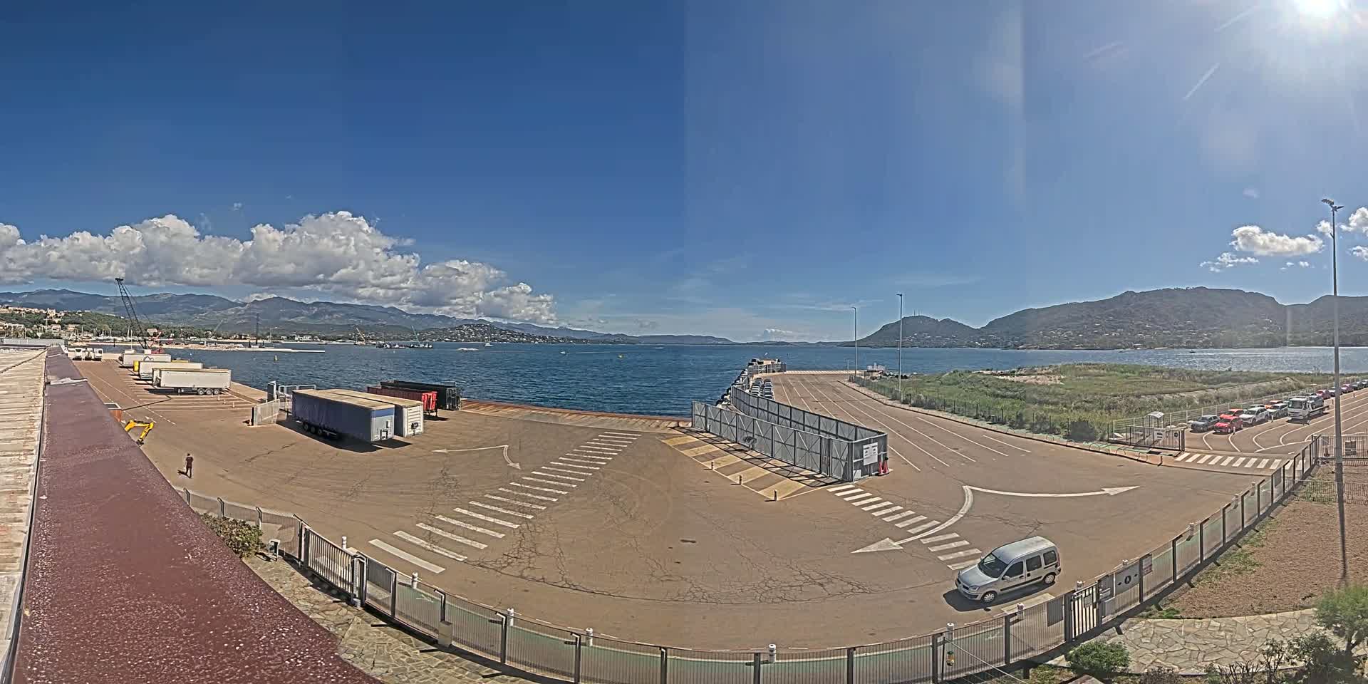 A sunny day shows a port area with trucks and cars parked, a body of water with mountains in the background, and a mostly empty paved lot with crosswalks.
