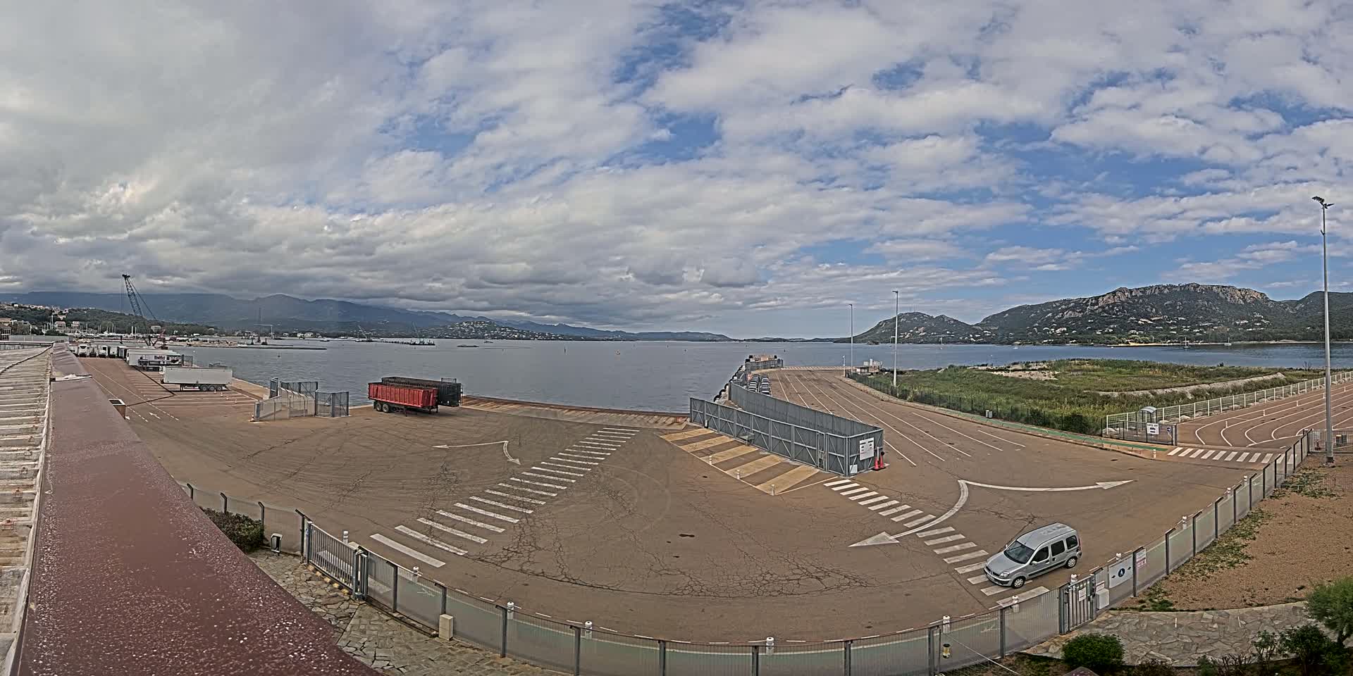 An empty port area with a few vehicles, under a partly cloudy sky, overlooks a calm body of water and distant mountains.