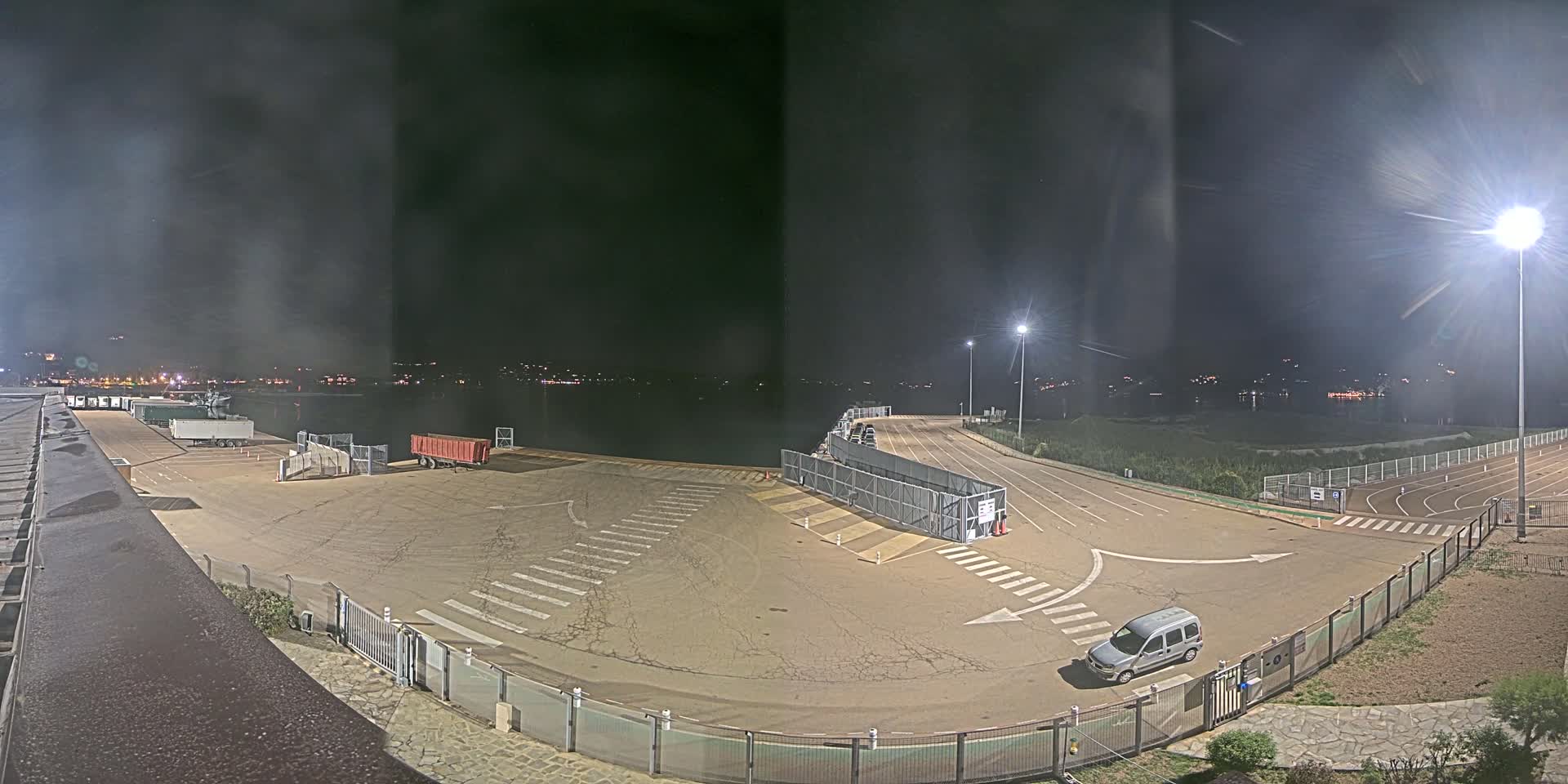 An empty nighttime port area with a few vehicles, illuminated by bright lights, overlooks a dark body of water with distant city lights.