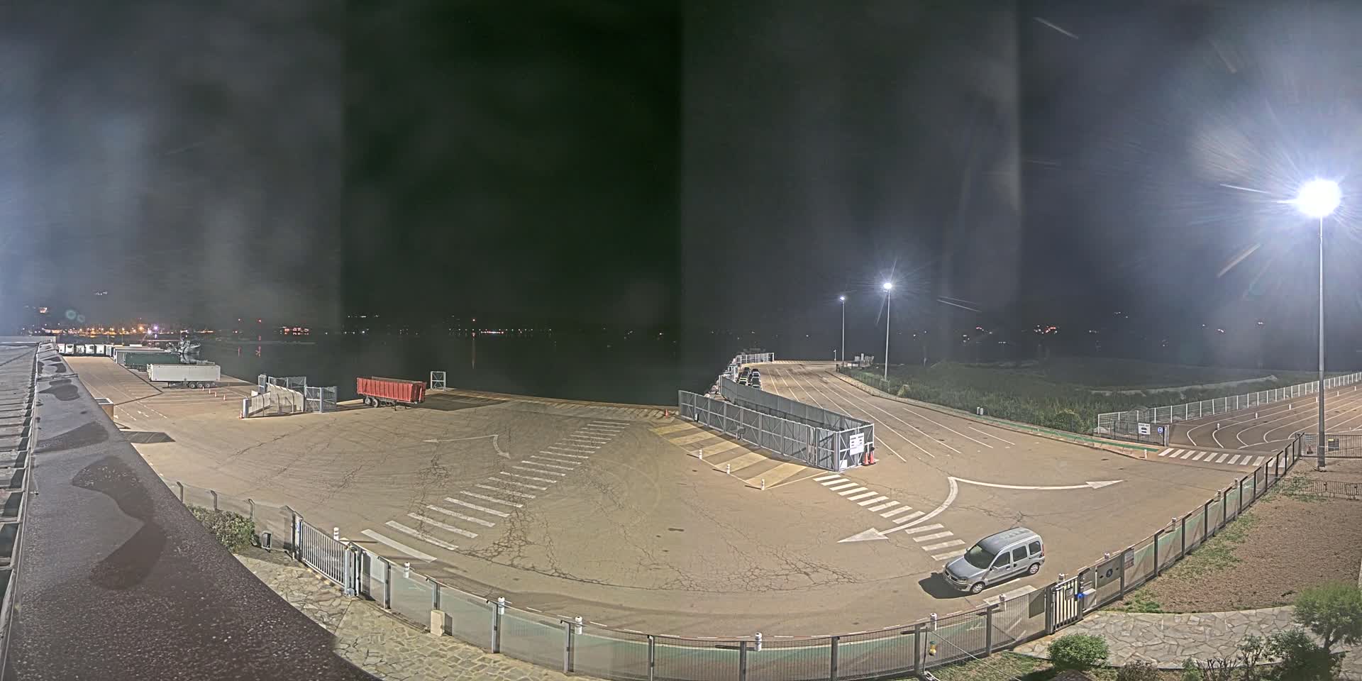 An empty nighttime port area with a single car and several trucks parked, illuminated by bright lights.