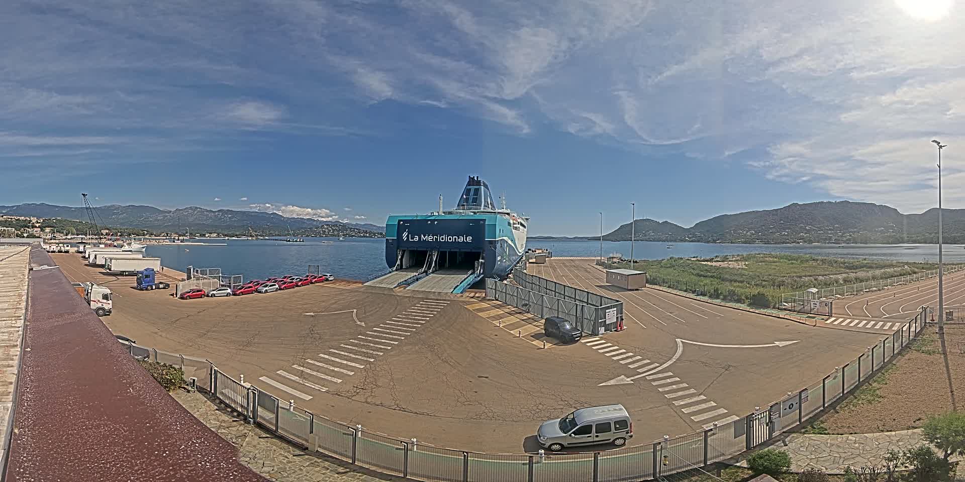 A large ferry is docked at a port on a sunny day with partly cloudy skies, surrounded by several vehicles and a few buildings.