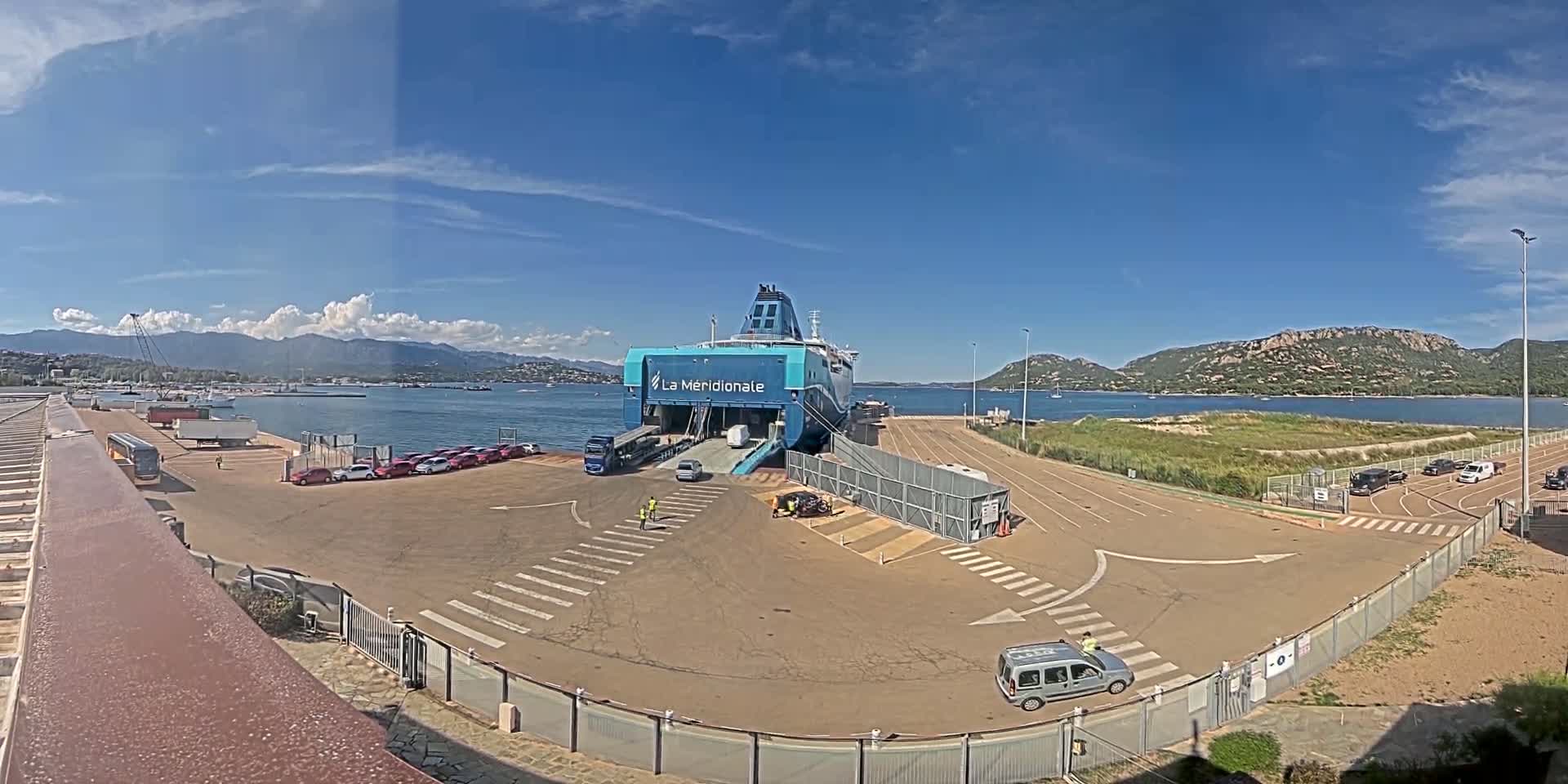 A large ferry is docked at a port on a sunny day, with several vehicles parked nearby and mountains visible in the distance.