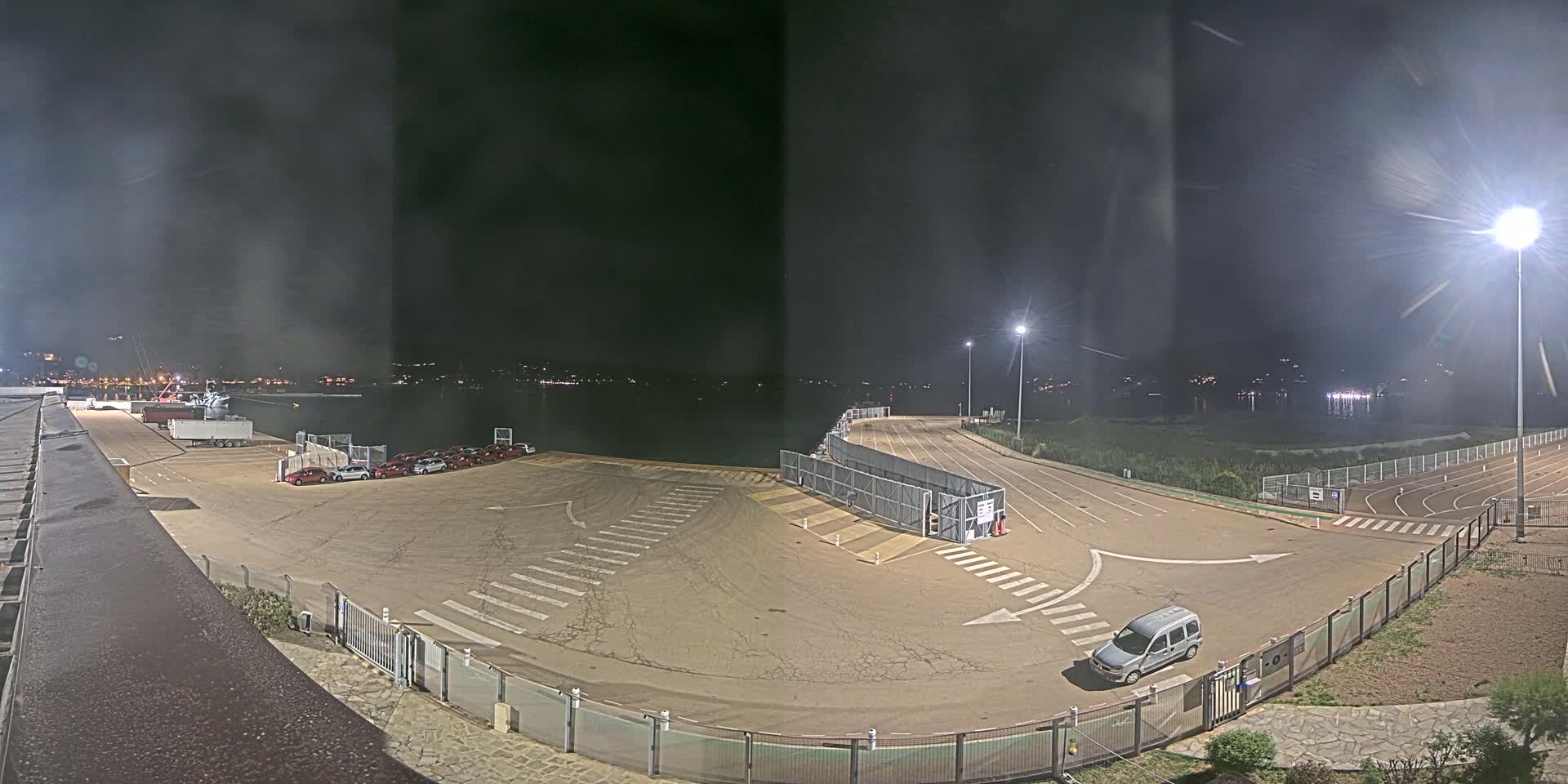 A nighttime view of an empty port area with a few parked cars, a van, and a body of water in the background under dark, hazy skies.