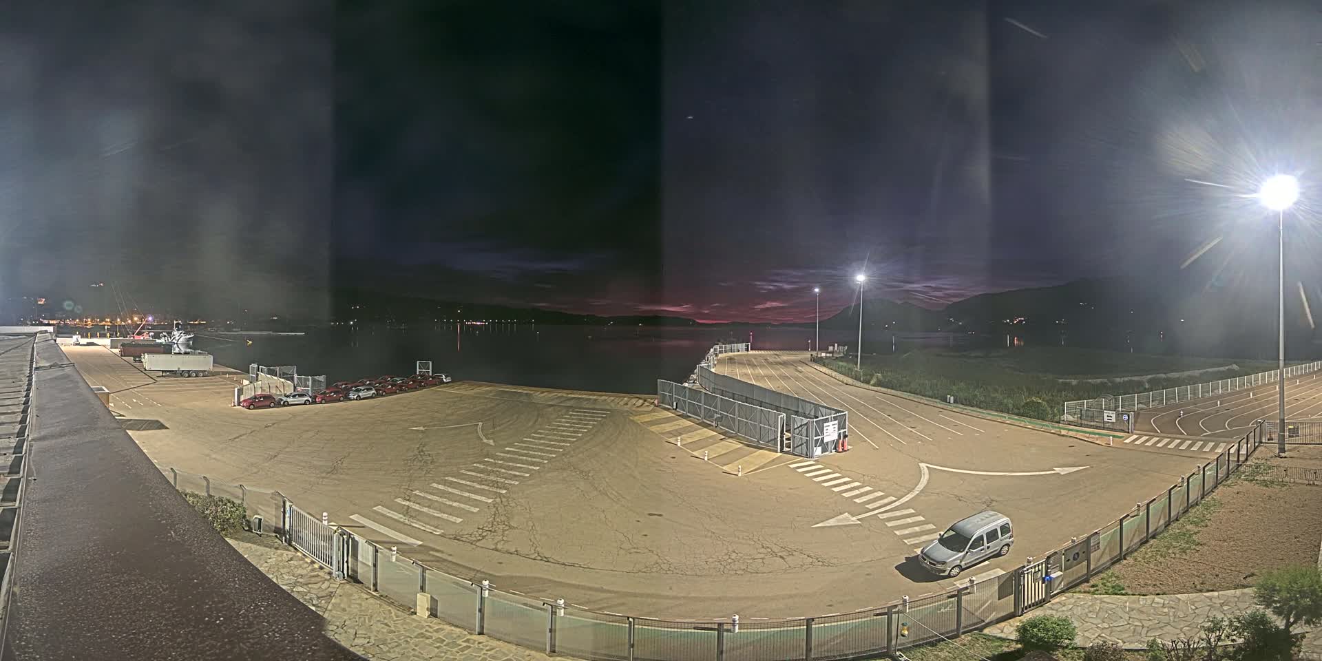 An empty, asphalt port area at night under a dusky sky, with a few vehicles parked and a body of water visible in the background.