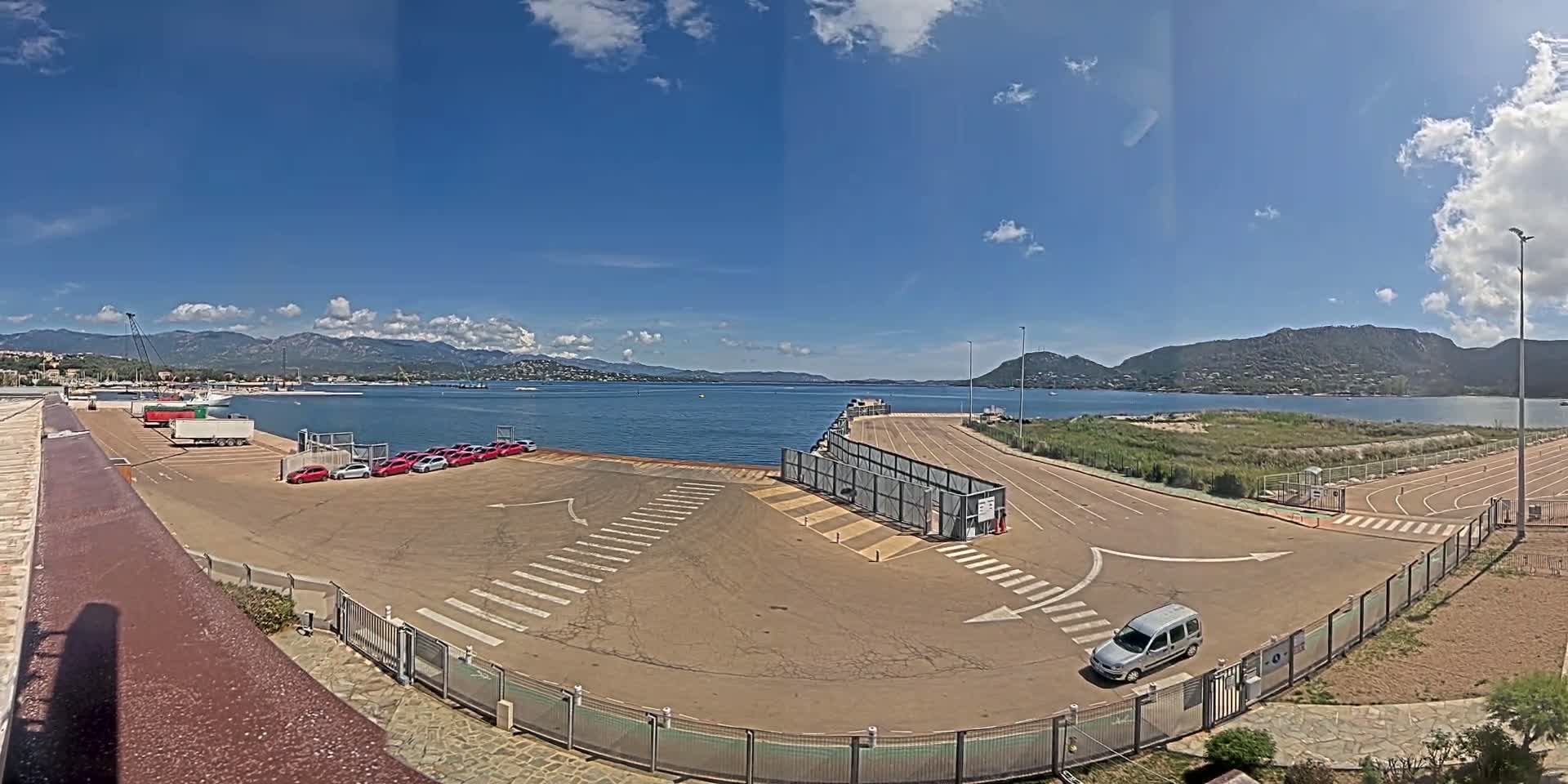 A mostly sunny day shows a harbor area with a parking lot containing several red cars and a lone silver van, near a body of water with mountains in the distance.