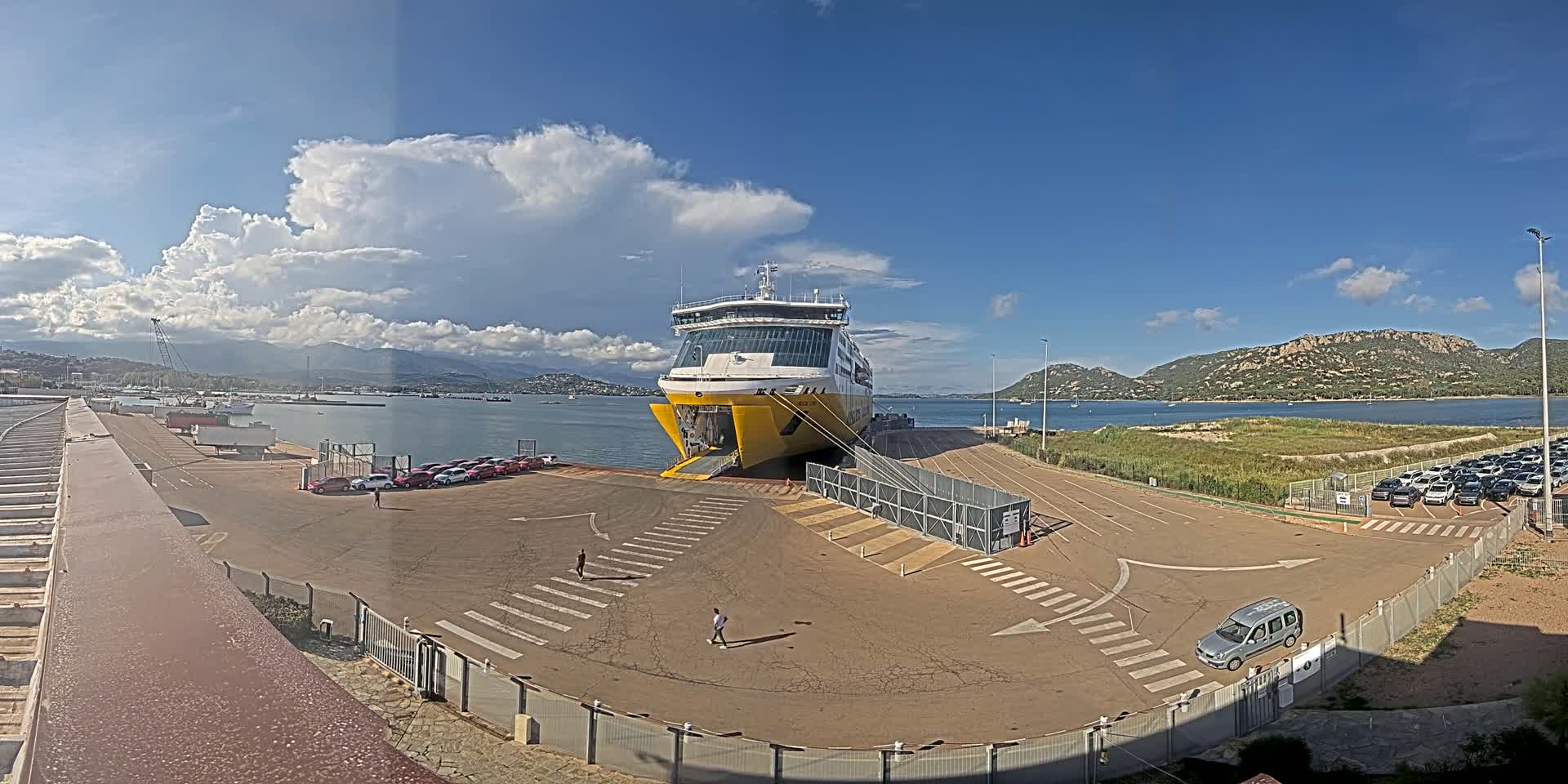 A large yellow and white ferry is docked at a port under a partly cloudy sky, with several cars parked nearby and mountains visible in the distance.