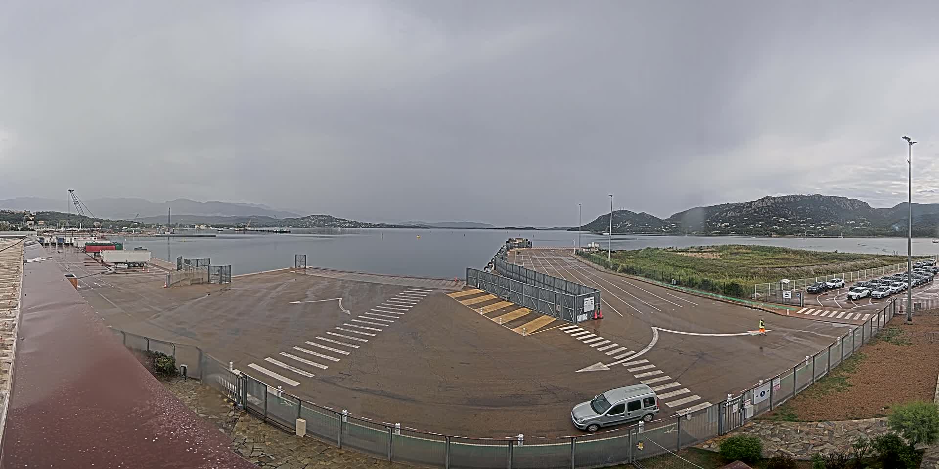 A wet, overcast day at a ferry terminal shows a mostly empty parking lot, a calm body of water beyond a gated area, and mountains in the distance.