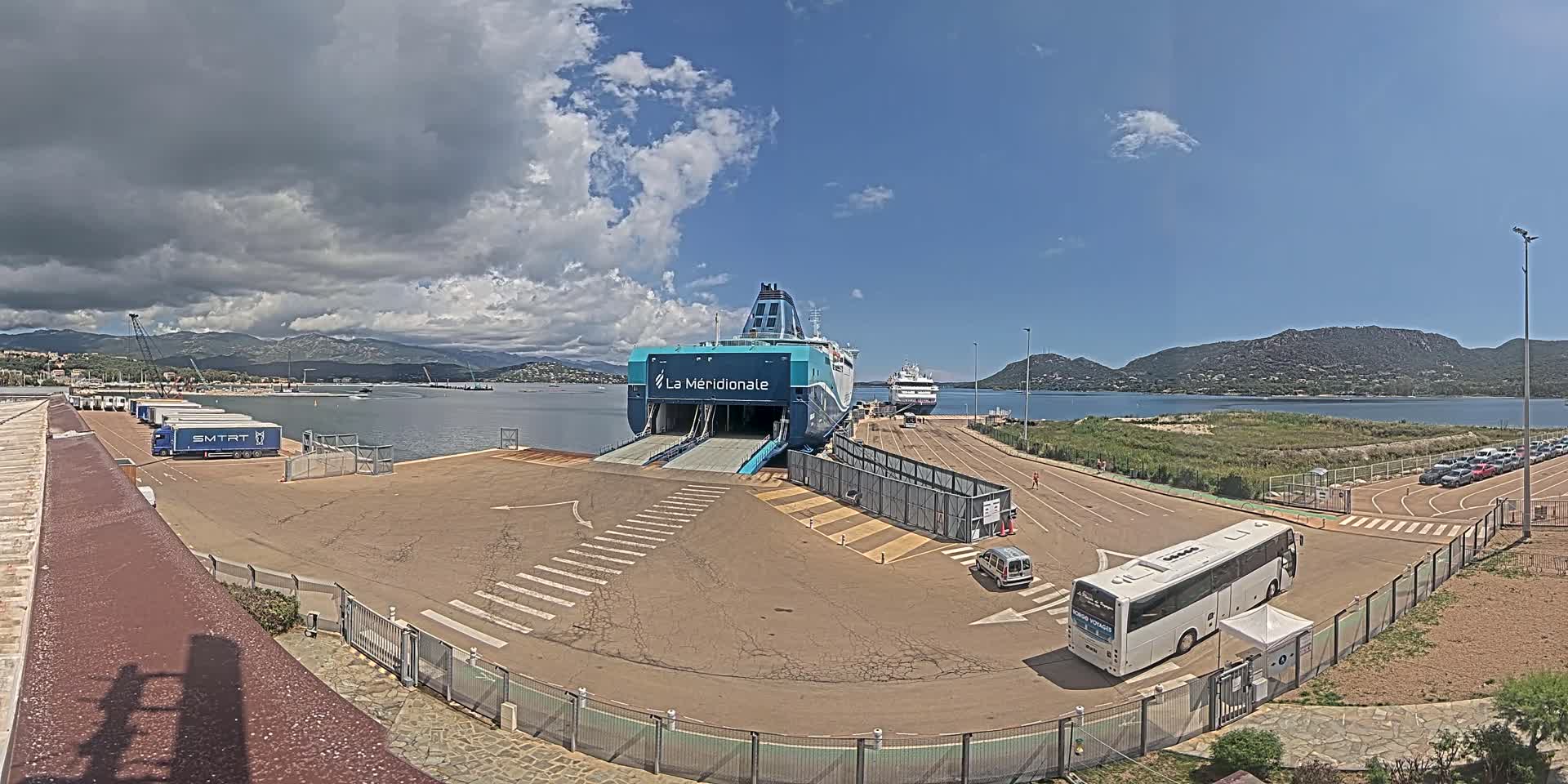 A ferry is docked at a port under partly cloudy skies, with several vehicles and a bus nearby.