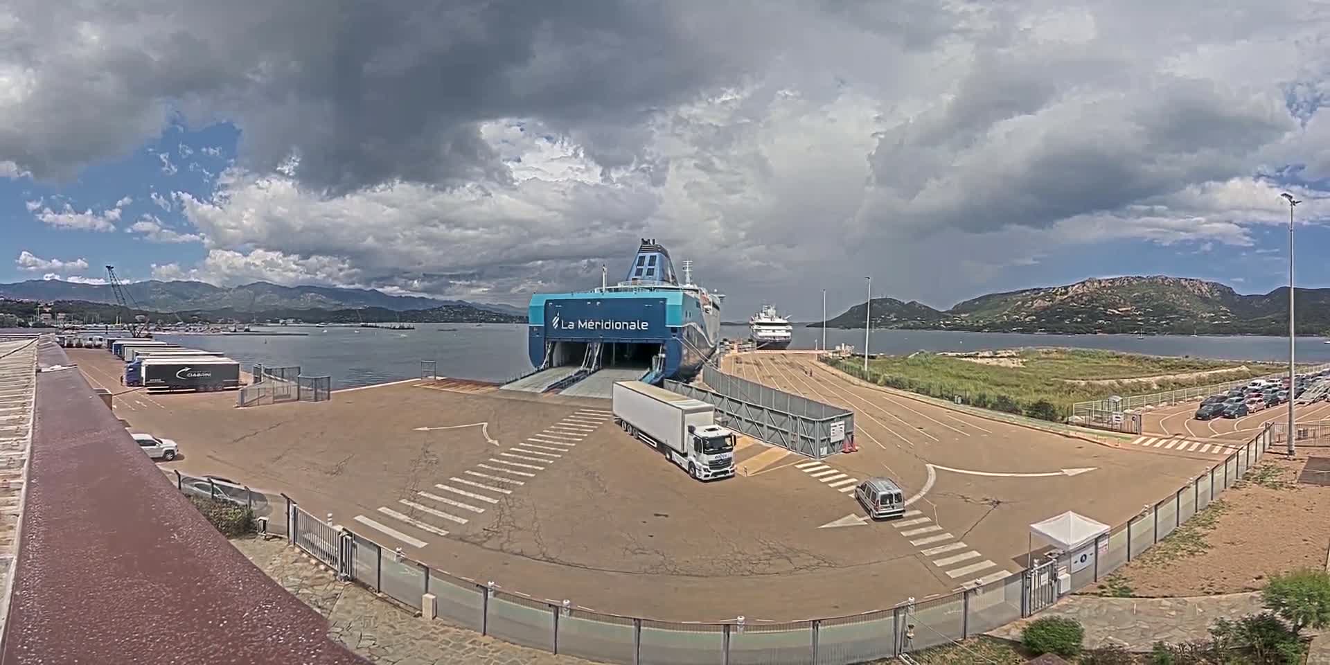 A large ferry is docked at a port under a partly cloudy sky, with trucks and cars in the surrounding area.