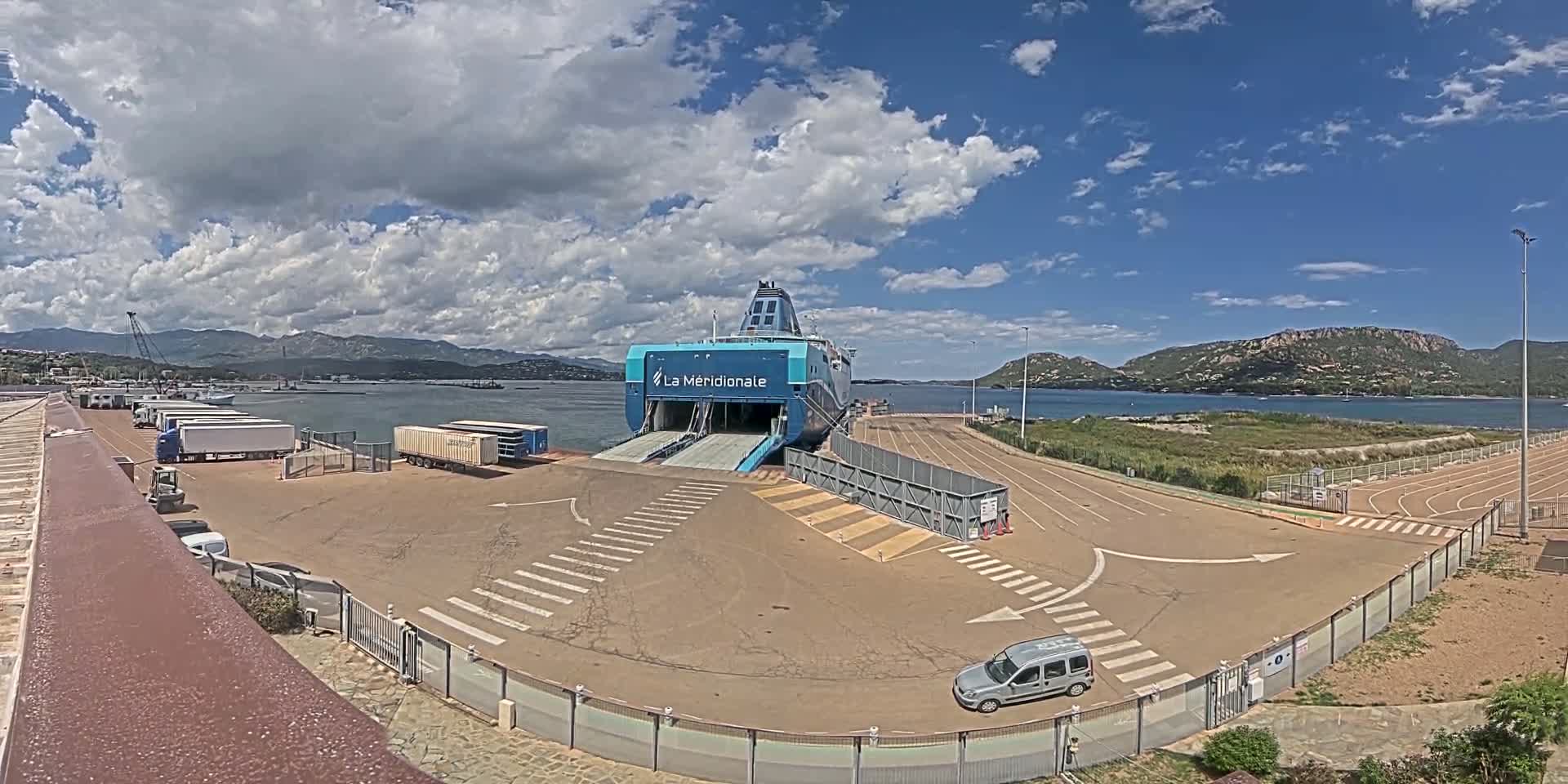 A large ferry is docked at a port on a partly cloudy day, with several trucks and a car nearby.