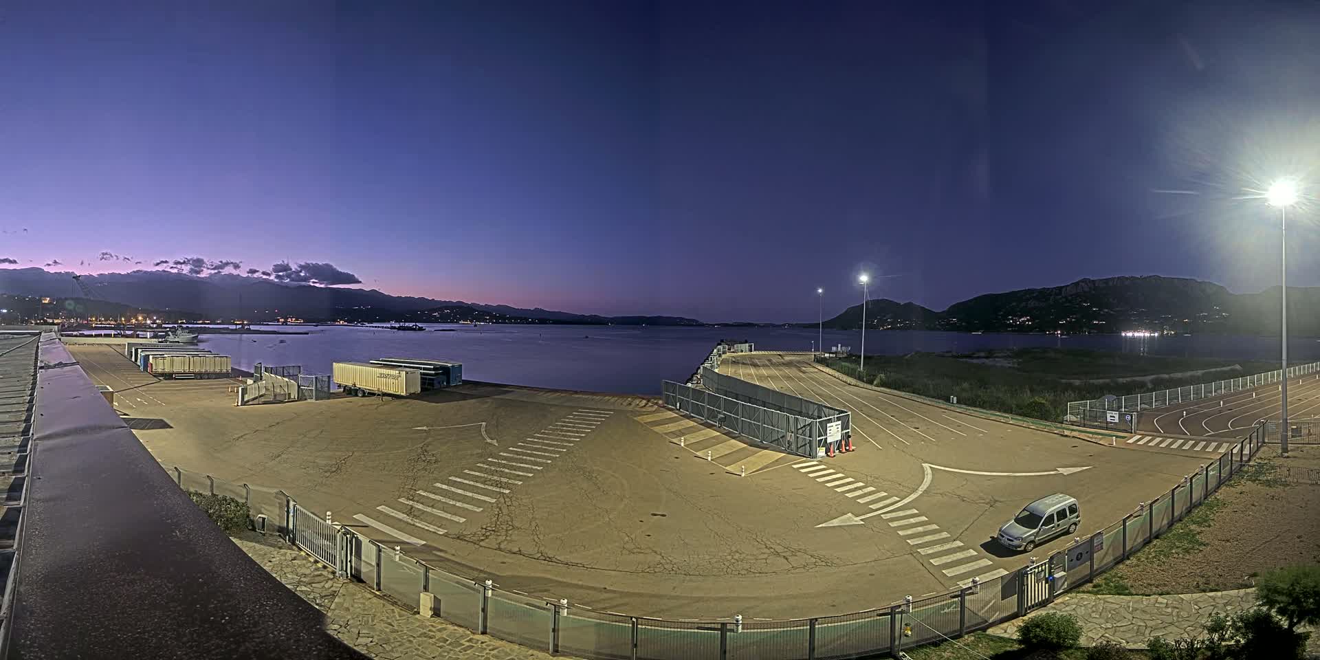 An empty, well-lit port area at twilight overlooks calm water and distant mountains under a clear, purple-tinged sky.