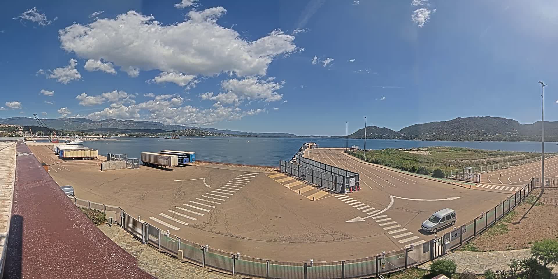 A ferry terminal with several cargo containers and a single vehicle in a mostly empty parking lot, overlooks calm water under a partly cloudy sky and mountains in the distance.
