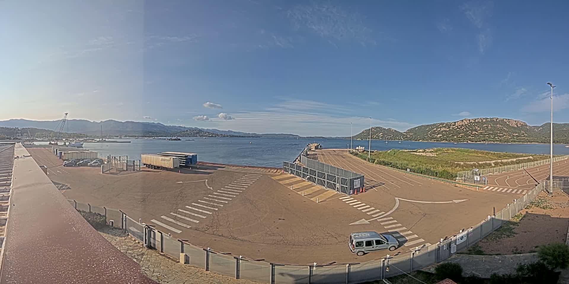 An empty port area with a few vehicles, under a mostly sunny sky, overlooks calm water and distant mountains.