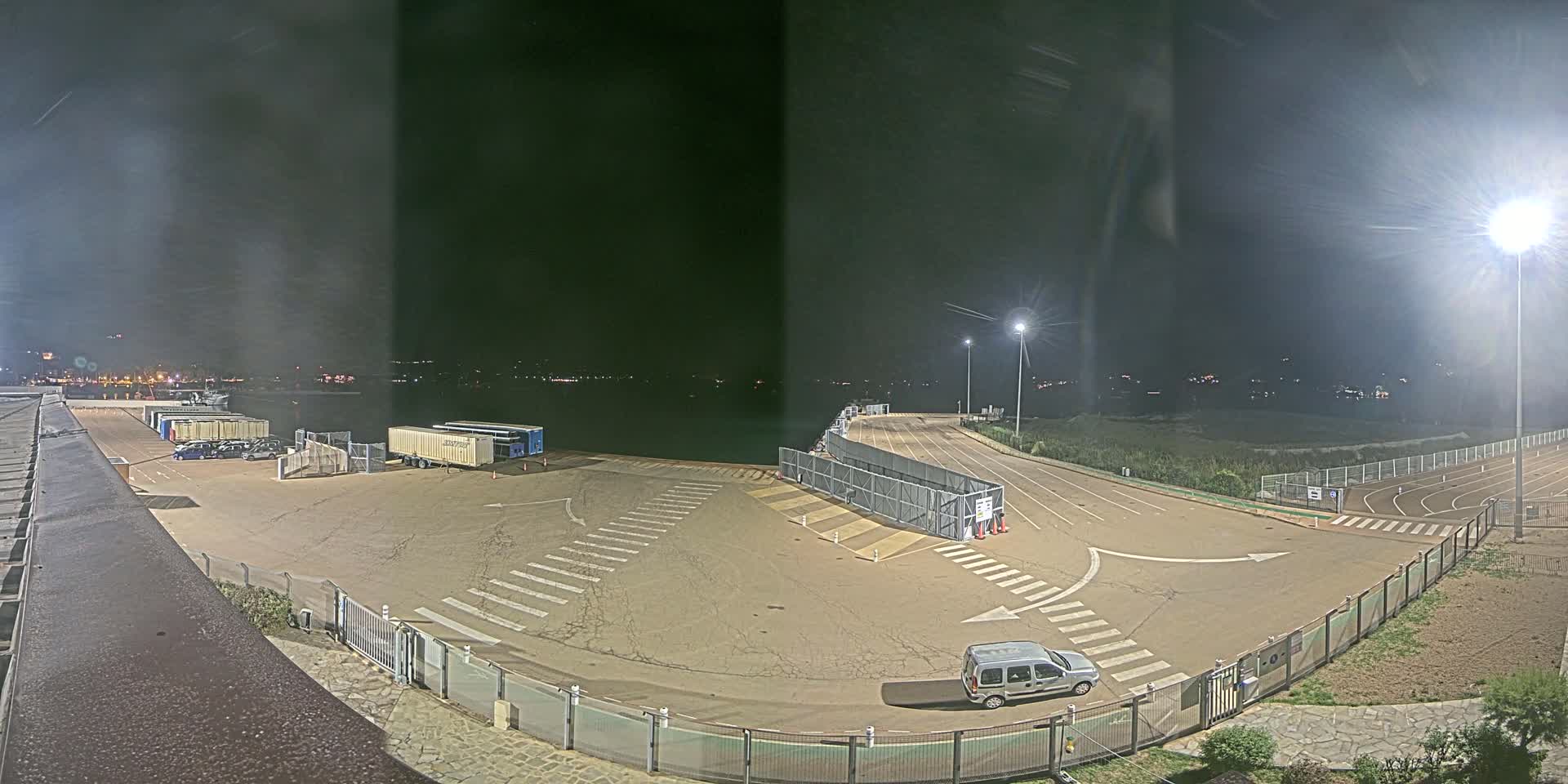 At night, an empty port area with a single vehicle is brightly lit, overlooking a dark body of water.