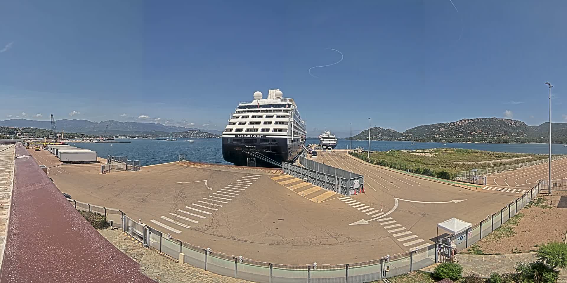 Two cruise ships are docked at a port on a sunny day, with mountains visible in the distance.