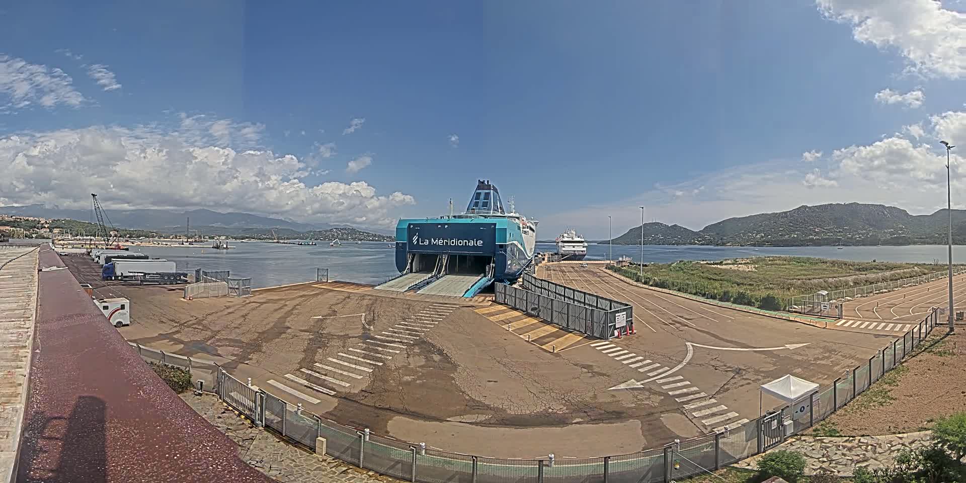 A large ferry is docked at a port on a partly cloudy day, with another smaller ship nearby and mountains in the background.