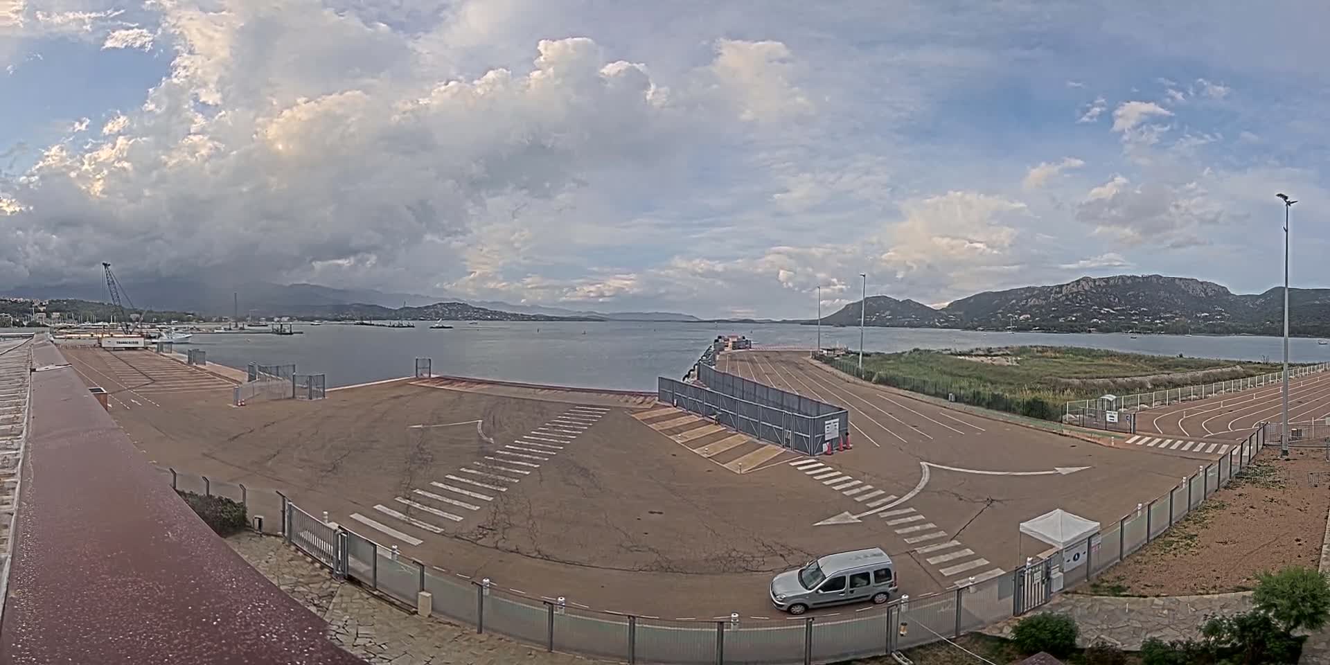 A mostly empty paved area with marked lanes and a single vehicle, near a calm body of water under a partly cloudy sky, with mountains visible in the distance.