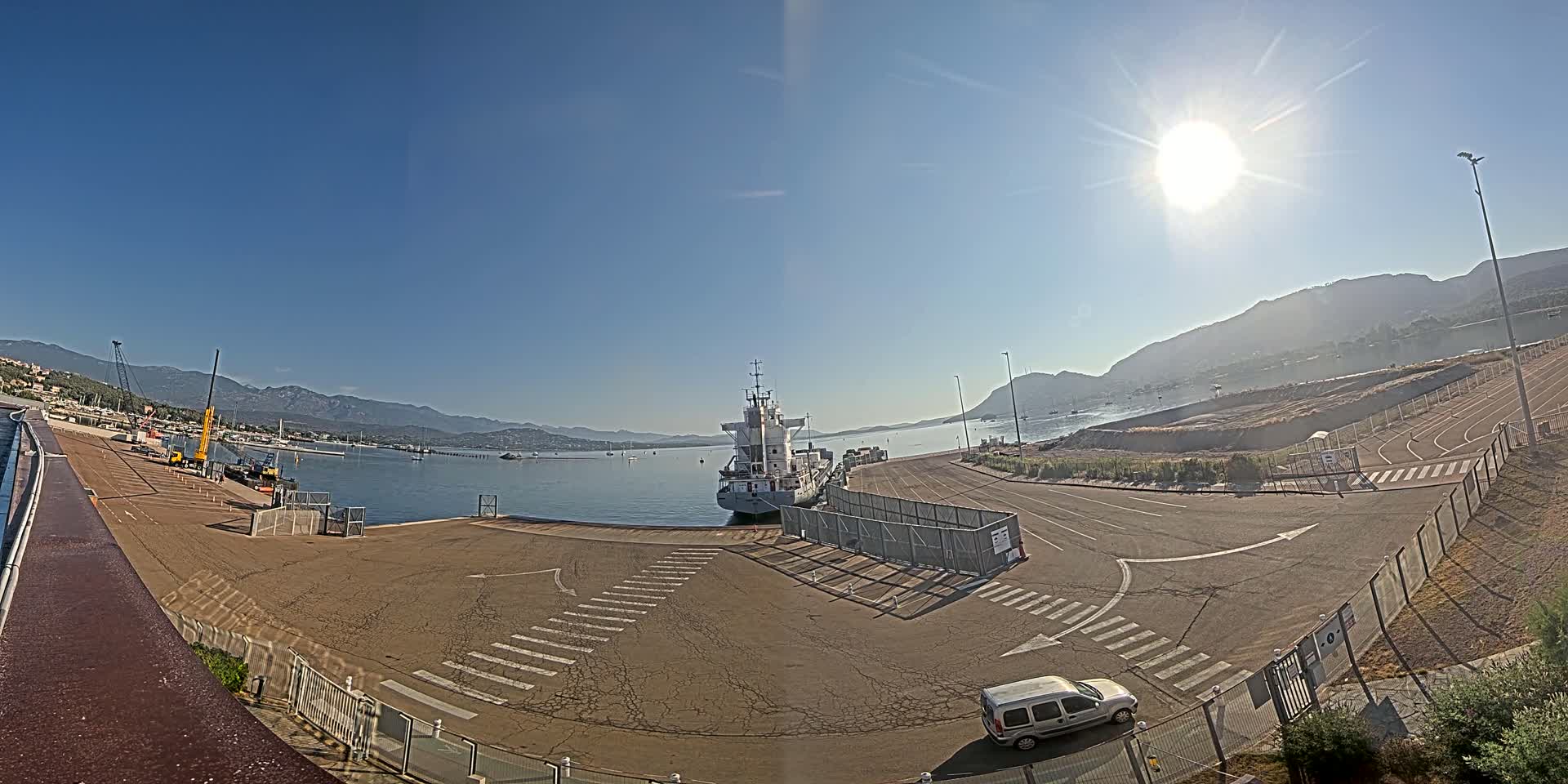 A cargo ship is docked at a port on a sunny day, with mountains in the background and a few other boats visible in the calm water.