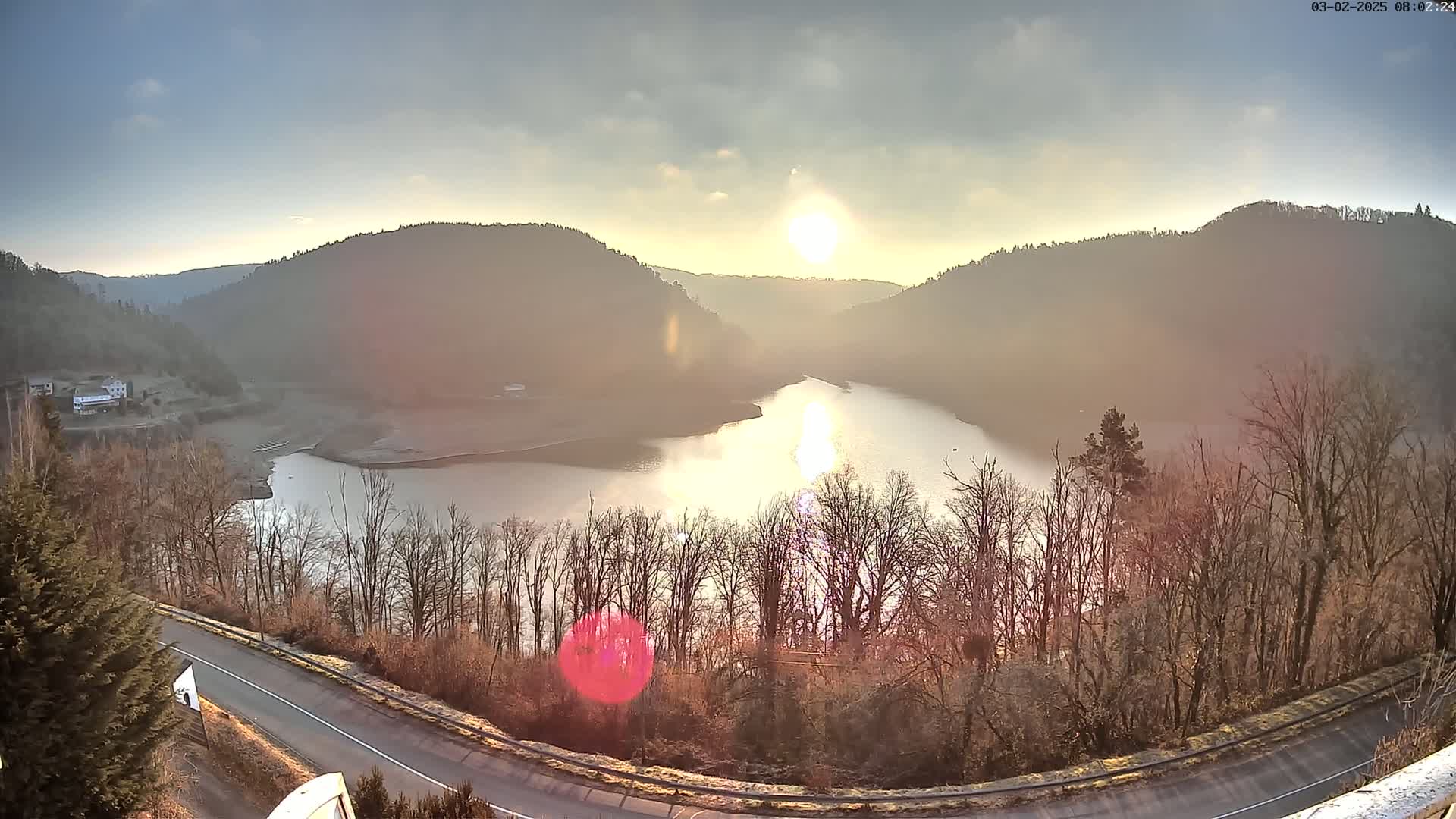 A sunlit lake nestled between hills, partially shrouded in a hazy atmosphere, is viewed from above a curving road.