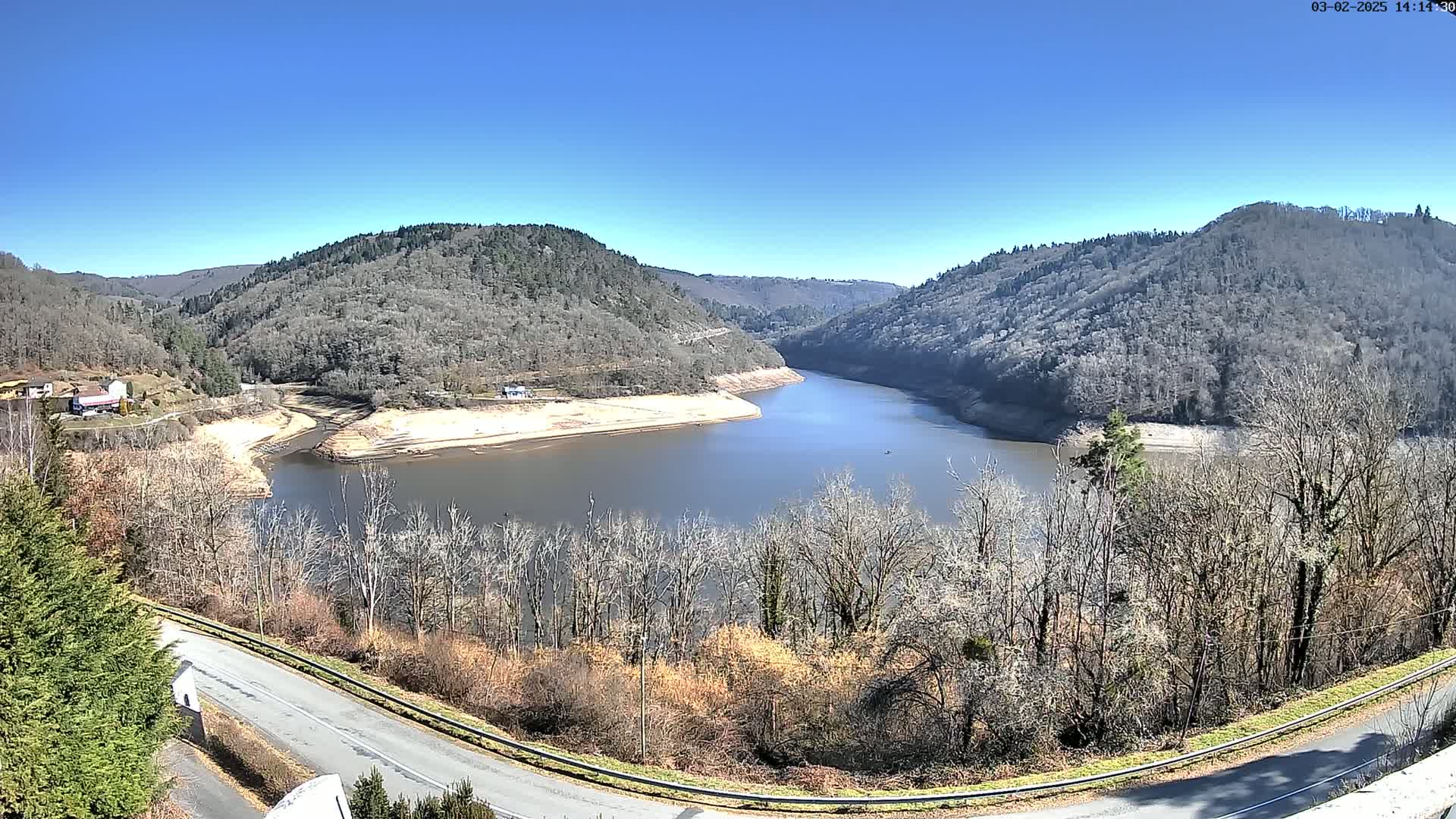 A winding river flows through a valley, surrounded by bare, winter trees and hills under a clear, sunny sky.