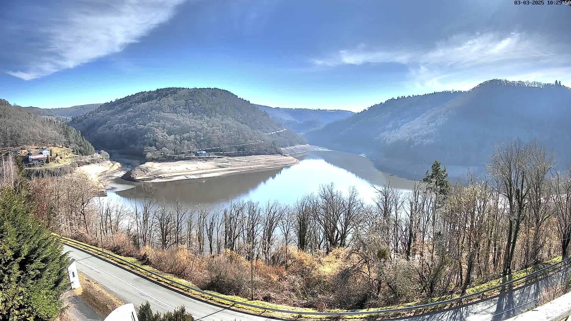 A partly sunny day reveals a winding reservoir nestled between tree-covered hills, with a road curving in the foreground.