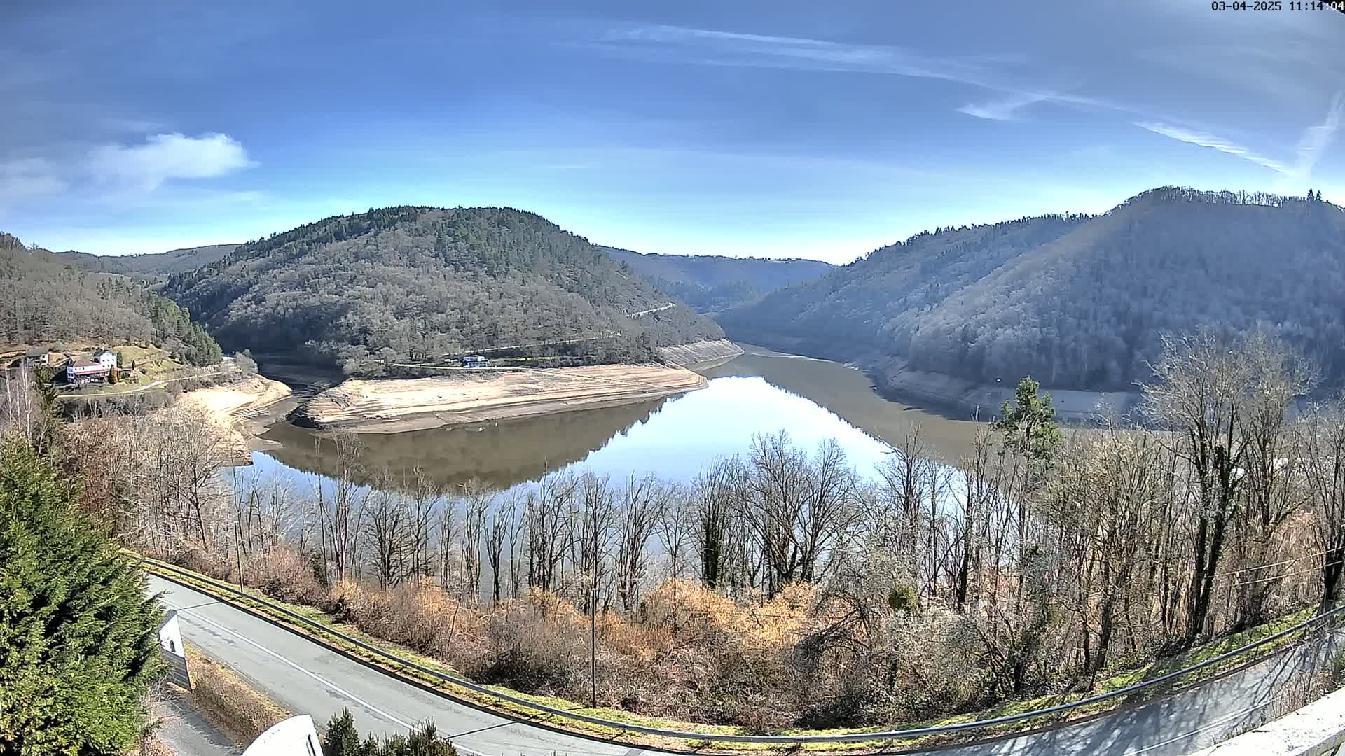 A partly sunny day reveals a calm, low-water reservoir nestled in a valley between barren, tree-covered hills, with a road curving along the lower edge of the view.